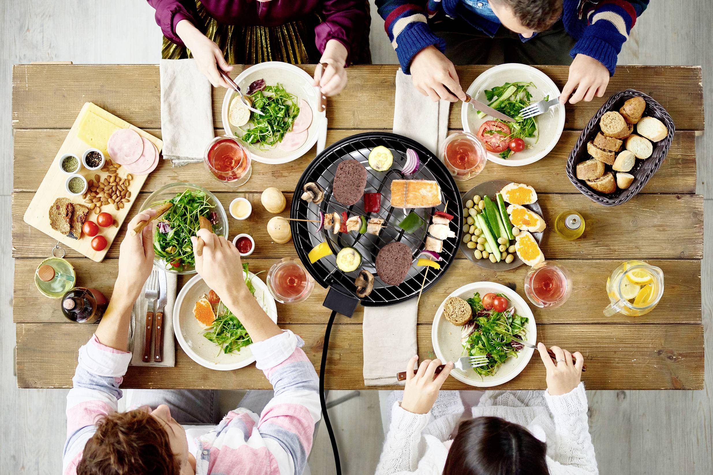 Fünf Personen sitzen an einem rustikalen Holztisch und genießen ein Essen mit Grillplatten, Salaten und Brot.