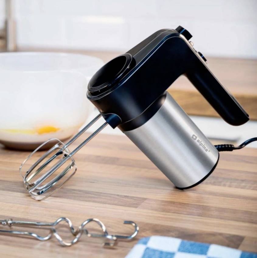 Ein silber-schwarzer Handmixer liegt auf einem Holztresen. Im Hintergrund steht eine Schüssel mit Eigelb. Ein Geschirrtuch ist daneben.