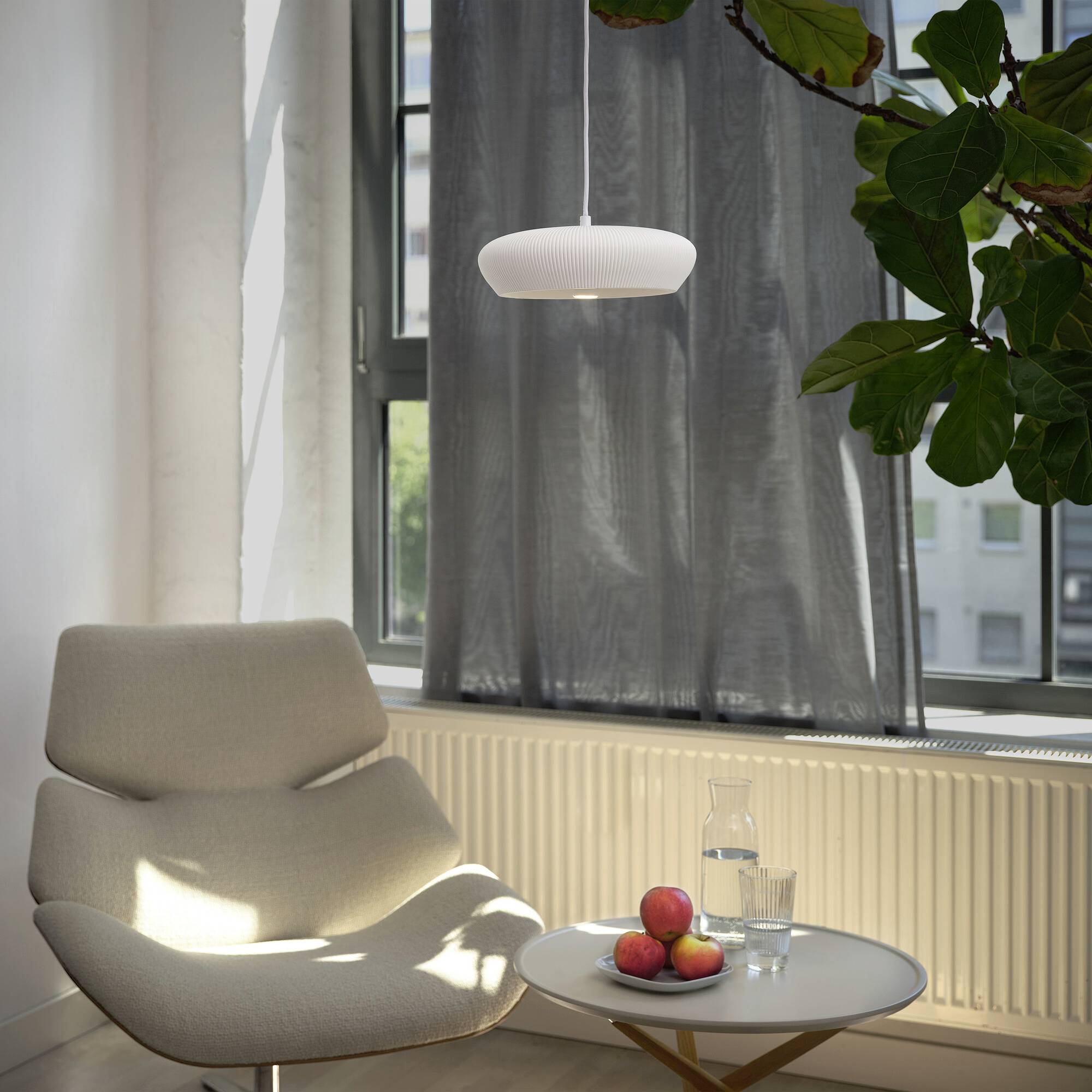 Ein moderner Sessel steht neben einem Tisch mit einem Glas Wasser und Äpfeln. Tageslicht fällt durch ein Fenster mit grauem Vorhang.
