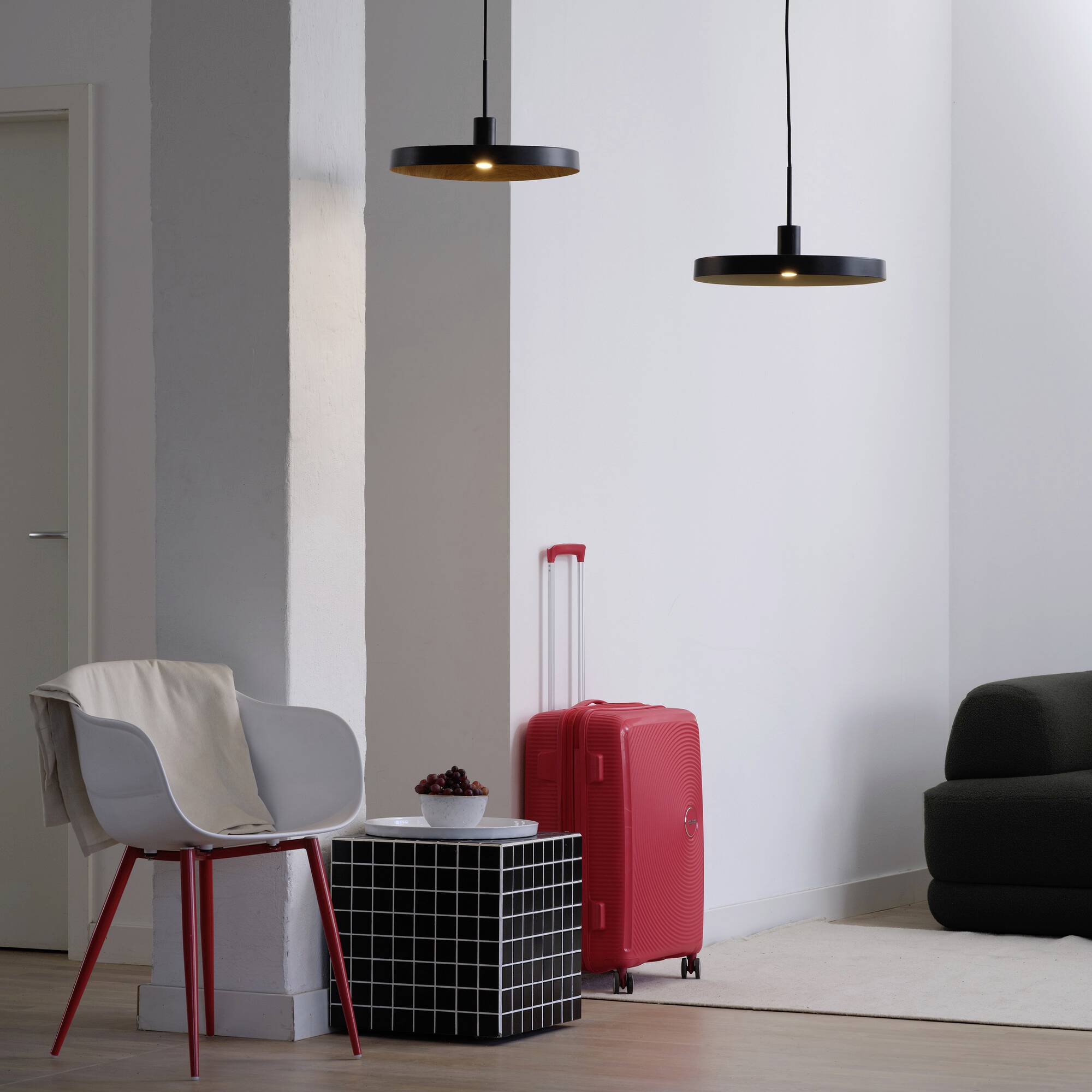 Ein modernes Wohnzimmer mit rotem Koffer neben schwarzem Beistelltisch, auf dem ein roter Helm liegt. Helle Wände und schwarze Pendelleuchten.