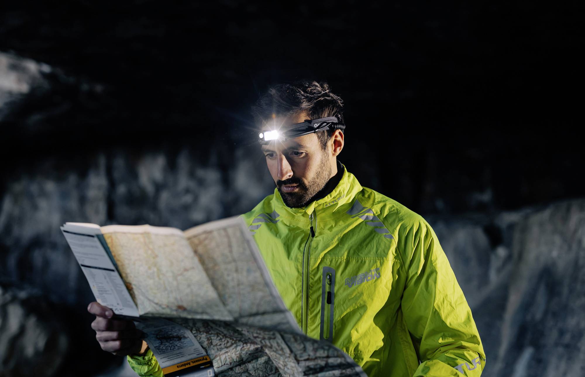 Ein Mann in einer reflektierenden Jacke liest mit Stirnlampe eine Karte in einer dunklen Höhle.