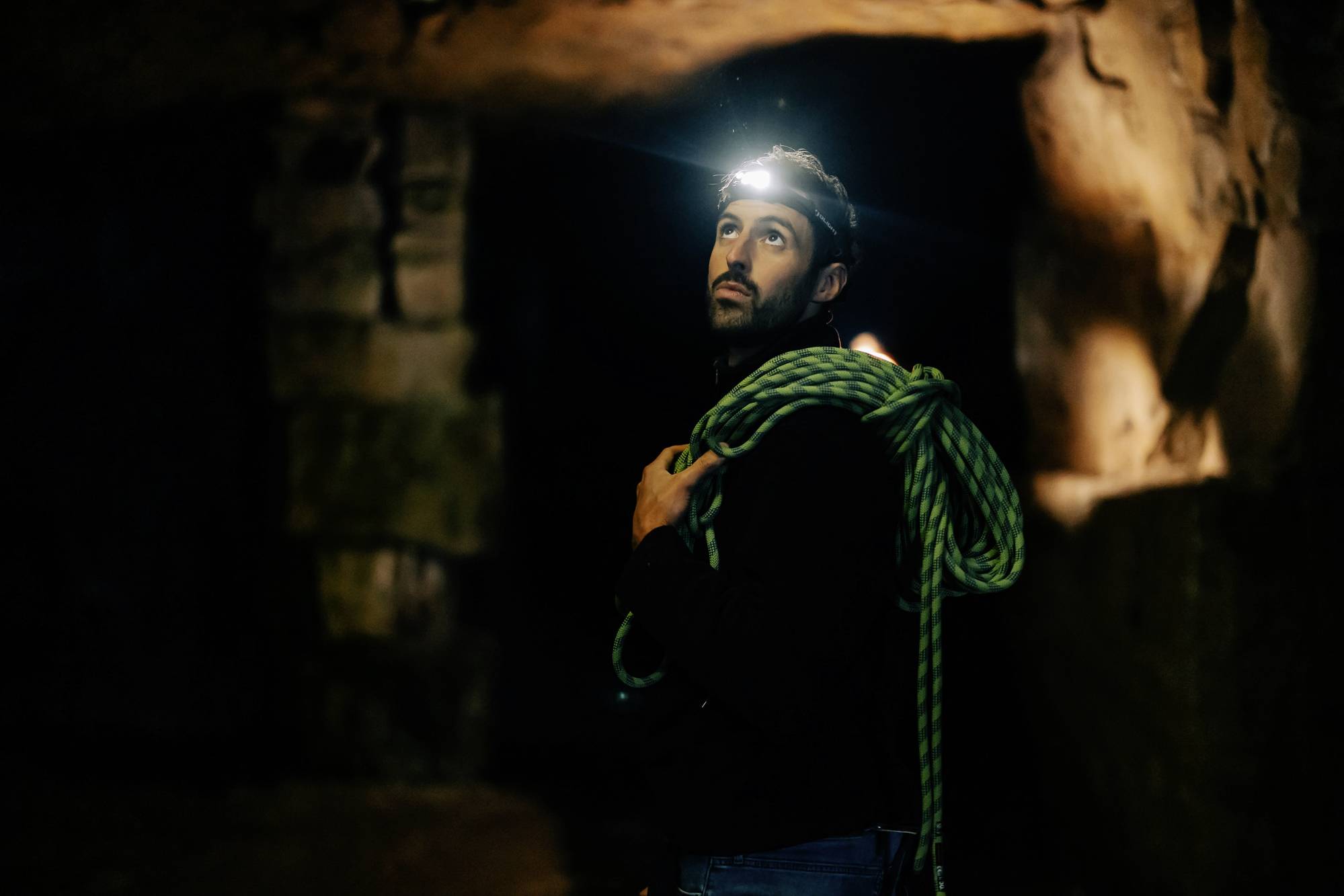Mann mit Stirnlampe und Kletterseil schaut in einer Höhle nach oben, von warmem Licht beleuchtet. Dunkle und rustikale Umgebung.