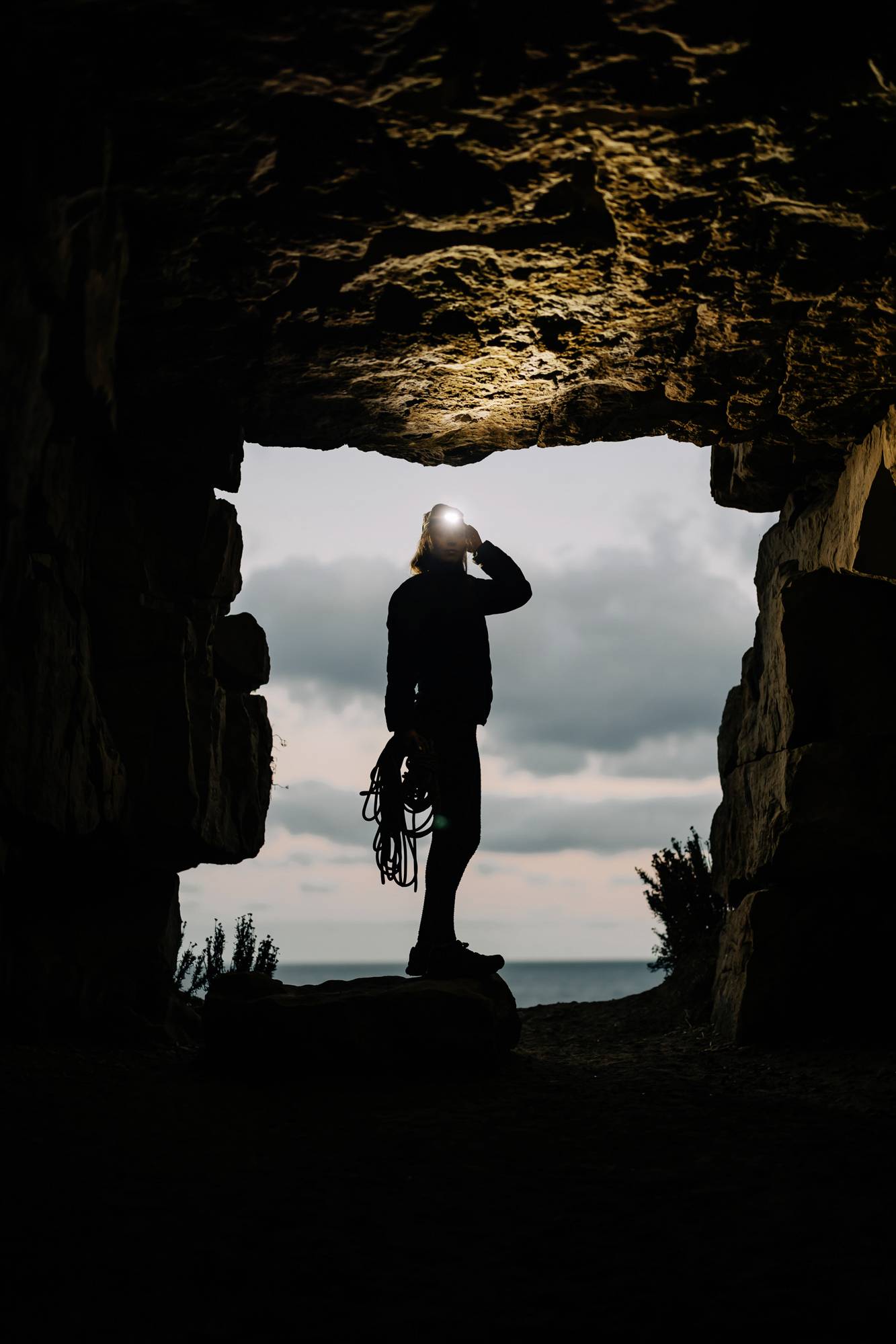 Eine Person mit Stirnlampe steht in einer Höhle und blickt hinaus auf das Meer bei Sonnenuntergang, hält Kletterseil in der Hand.