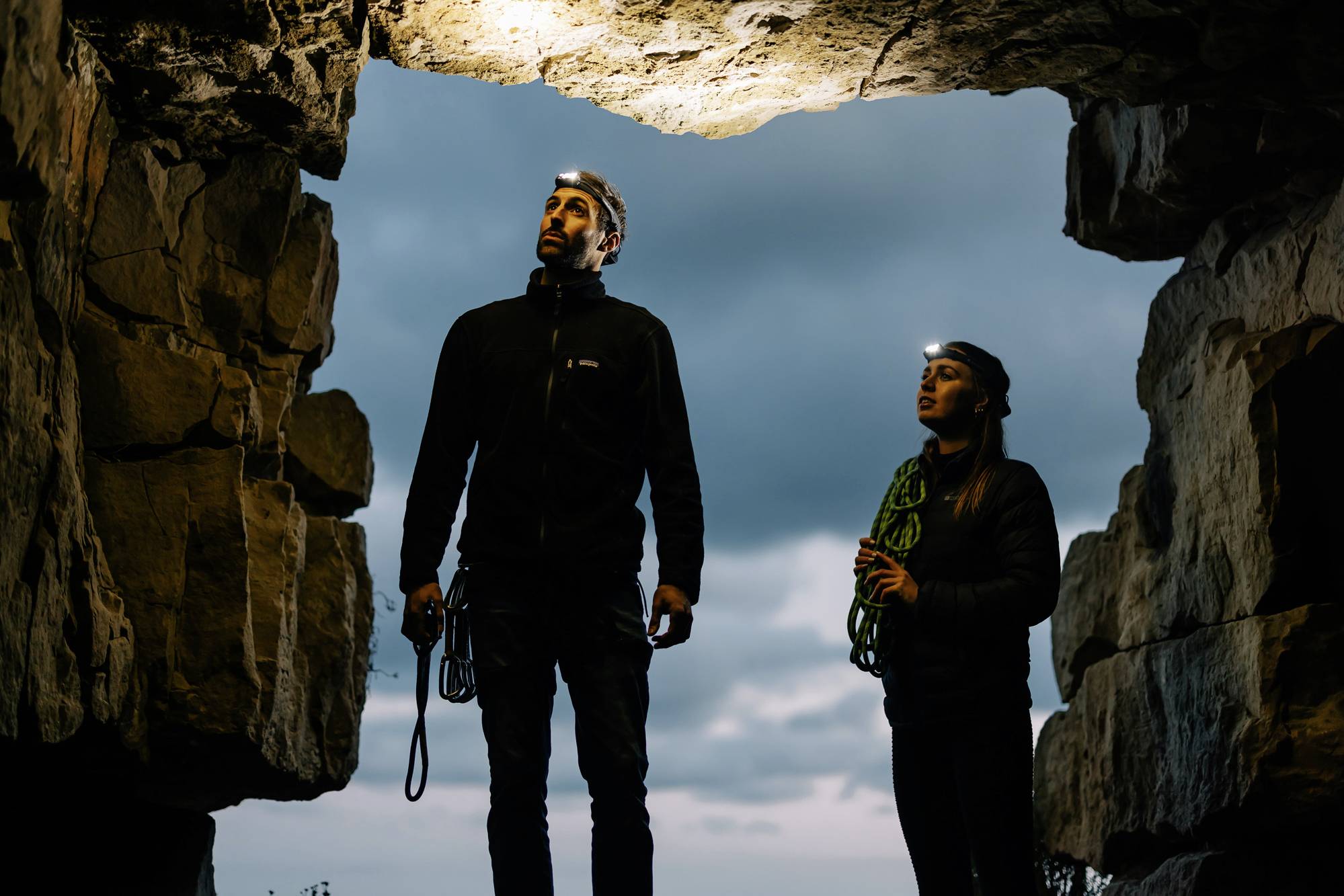 Zwei Personen mit Stirnlampen stehen in einer Felsöffnung im Freien. Sie schauen nach oben, umgeben von Felsen, bei bewölktem Himmel.
