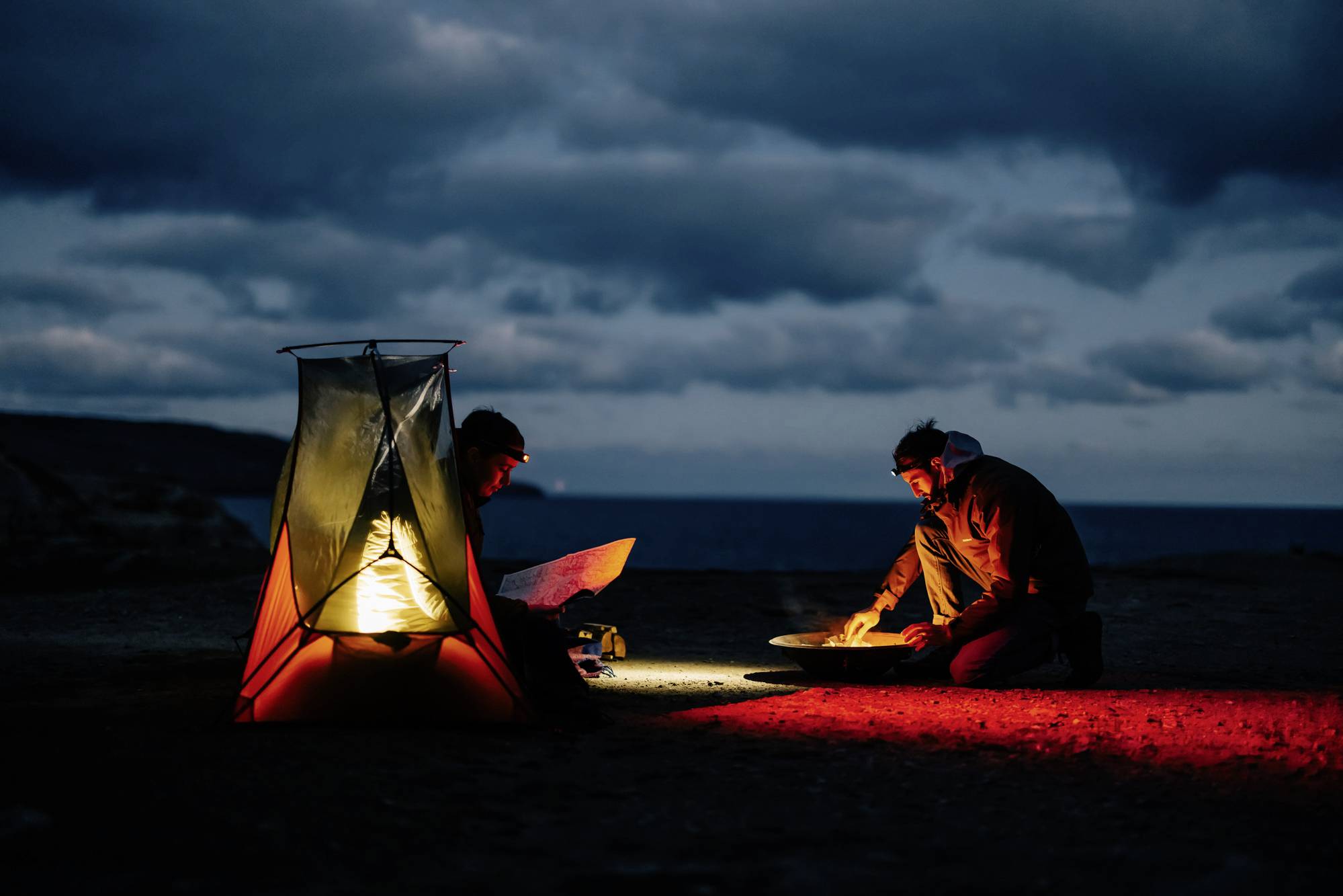 Zwei Personen campen bei Nacht am Meer, beleuchtet von einer Laterne. Eine liest eine Karte, die andere kocht über einem tragbaren Kocher. Bewölkter Himmel im Hintergrund.