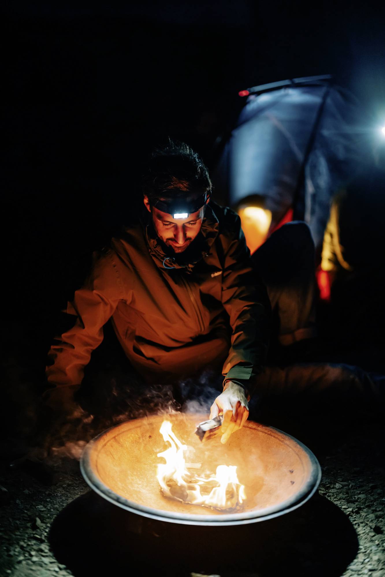 Ein Mensch sitzt nachts vor einer brennenden Feuerschale und trägt eine Stirnlampe. Im Hintergrund ist ein Zelt beleuchtet zu sehen.