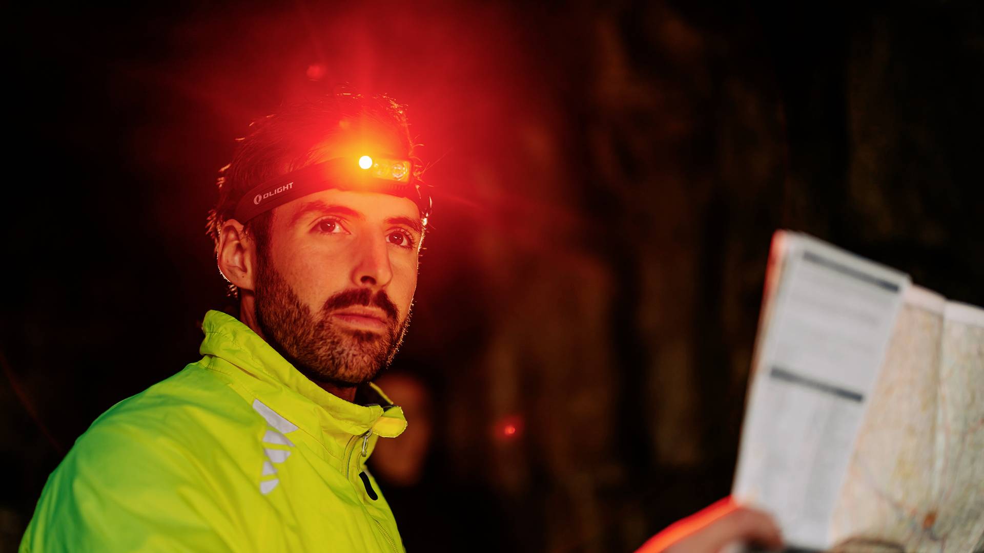 Ein Mann mit Stirnlampe hält eine Karte in der Hand. Er trägt eine auffällige, reflektierende Jacke, umgeben von einer dunklen Umgebung.