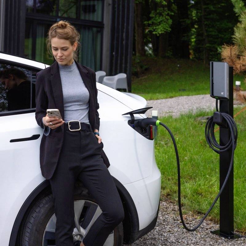 Person lehnt an einem Elektroauto, das an einer Ladestation angeschlossen ist; sie blickt auf ihr Handy in einem modernen Umfeld.