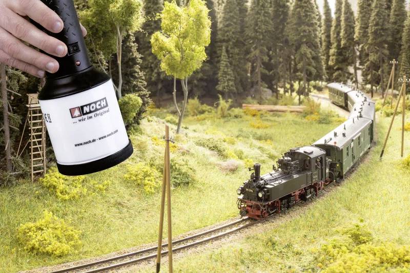 Ein kleines Modellzugmodell fährt durch eine üppige, bewaldete Landschaft, während eine Hand mit einer Lichtquelle darüber für die Szenerie-Verbesserung positioniert ist.