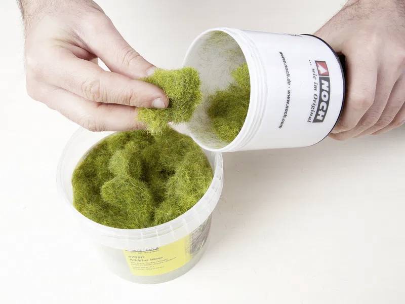 Eine Person schüttet grüne synthetische Grasfasern aus einem Behälter in eine größere Plastikwanne, die für Landschaftsmodelle oder Dioramen verwendet wird.