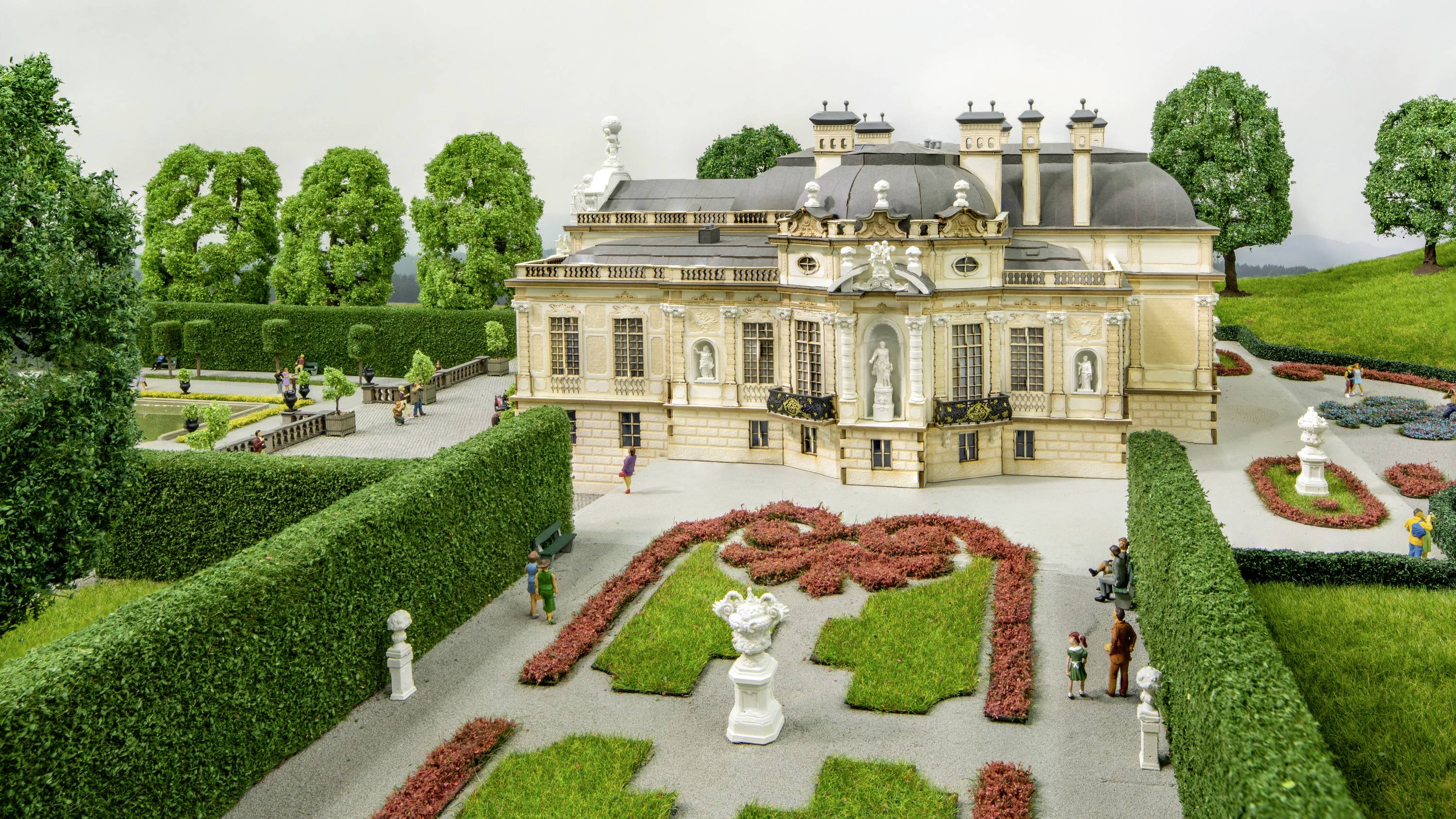 Ein Miniaturmodell eines prächtigen Schlosses mit gepflegten Gärten, Hecken und Statuen, umgeben von Bäumen und Spaziergängern.