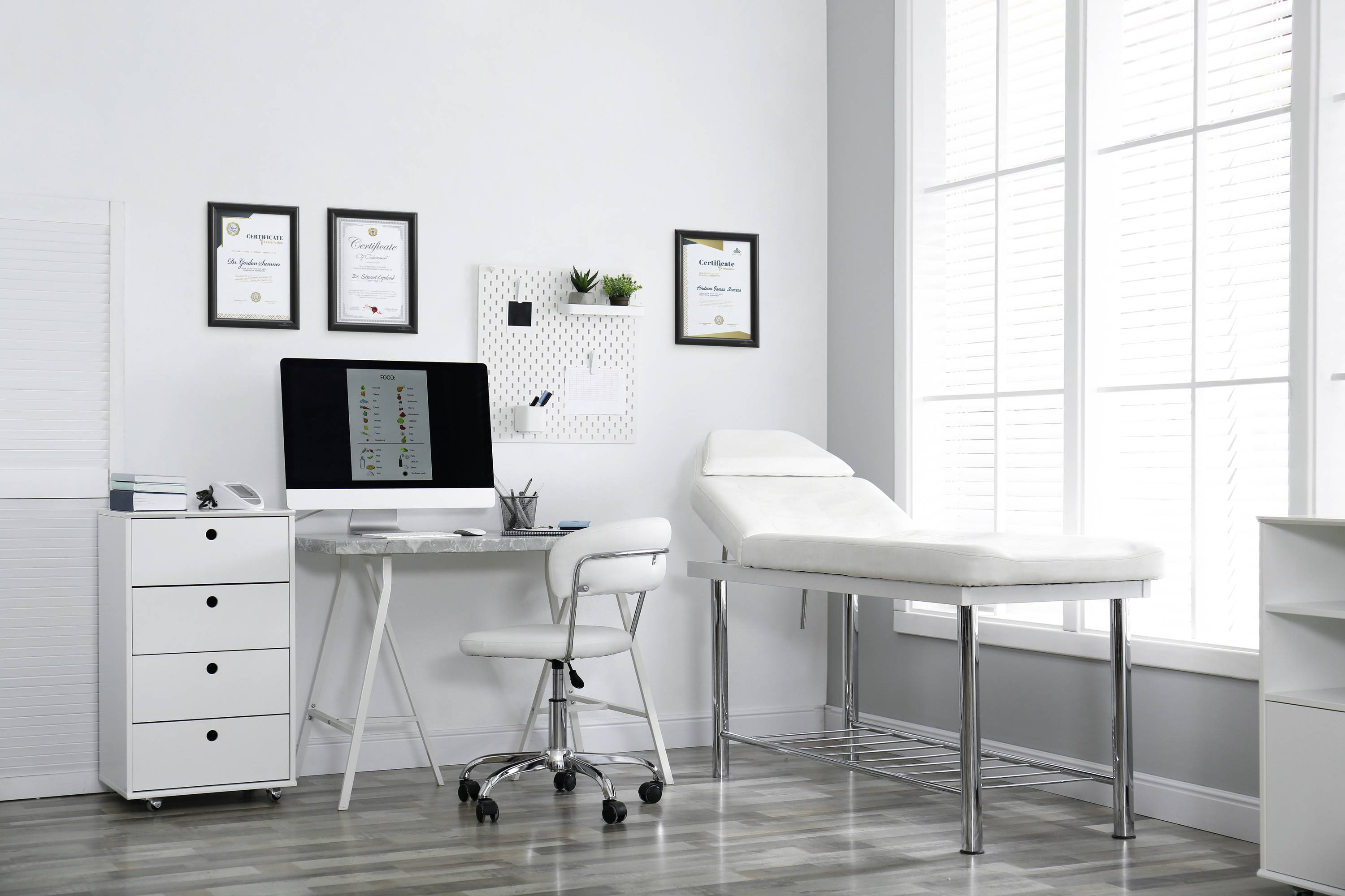 Moderner Arztpraxisraum mit Schreibtisch, Computer, Dokumenten an der Wand und Untersuchungsliege neben großen Fenstern.