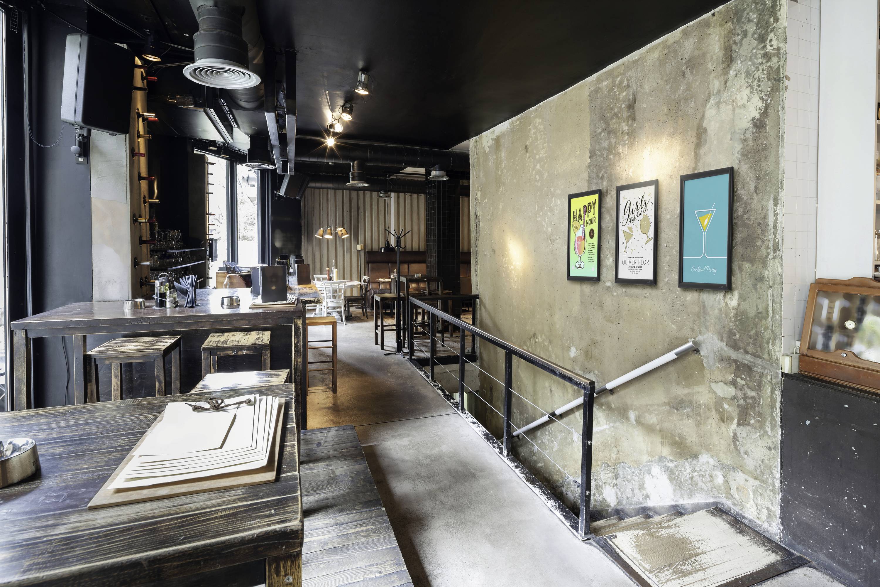 Ein modernes Café mit industriellem Design, Holztischen und Retro-Wandpostern auf rauen Wänden. Treppe führt zu einem unteren Bereich.