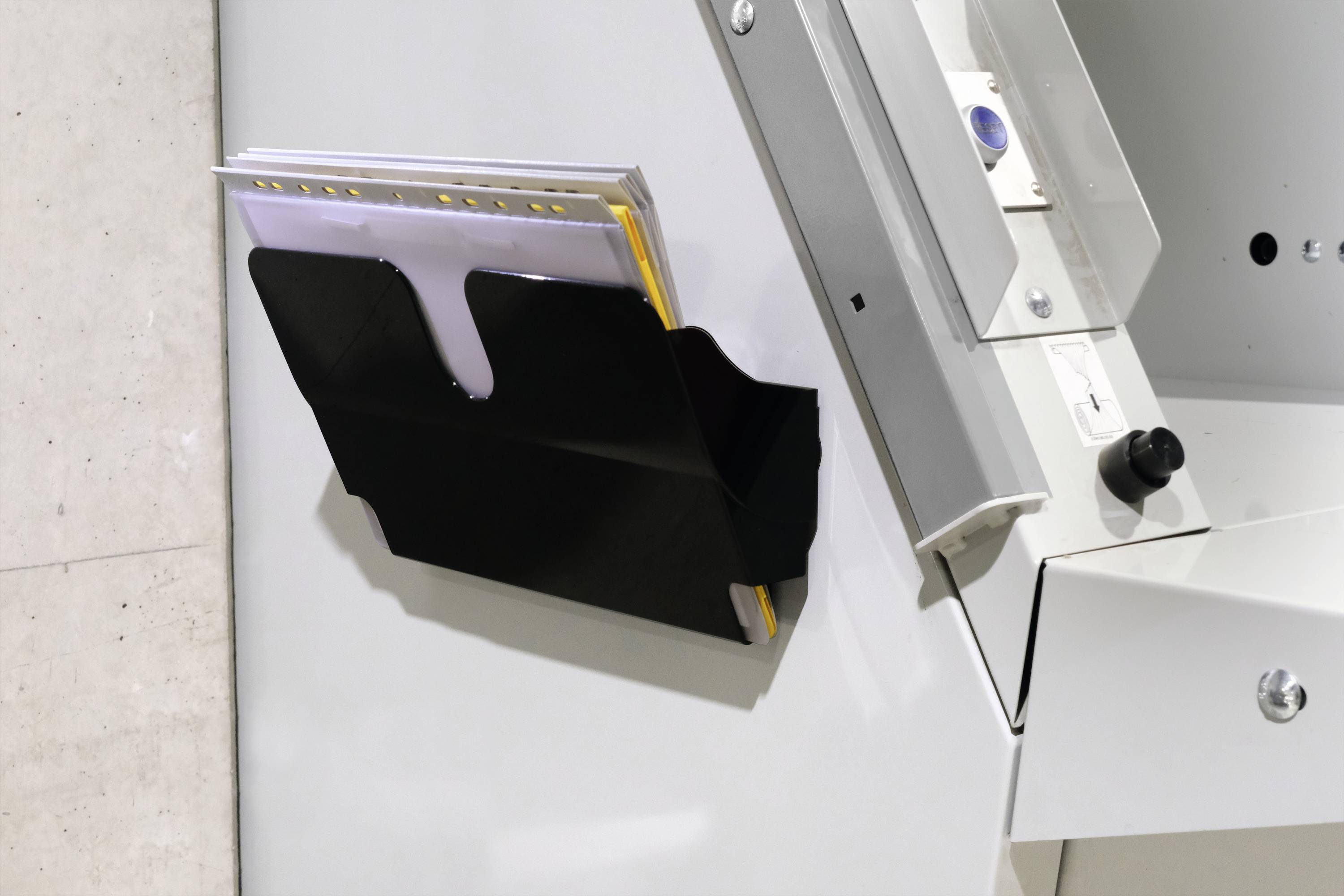 Schwarzer Wandhalter mit Akteneinsatz an einer weißen Wand; Halter enthält Hefte und Dokumente; minimalistisches Bürodesign.