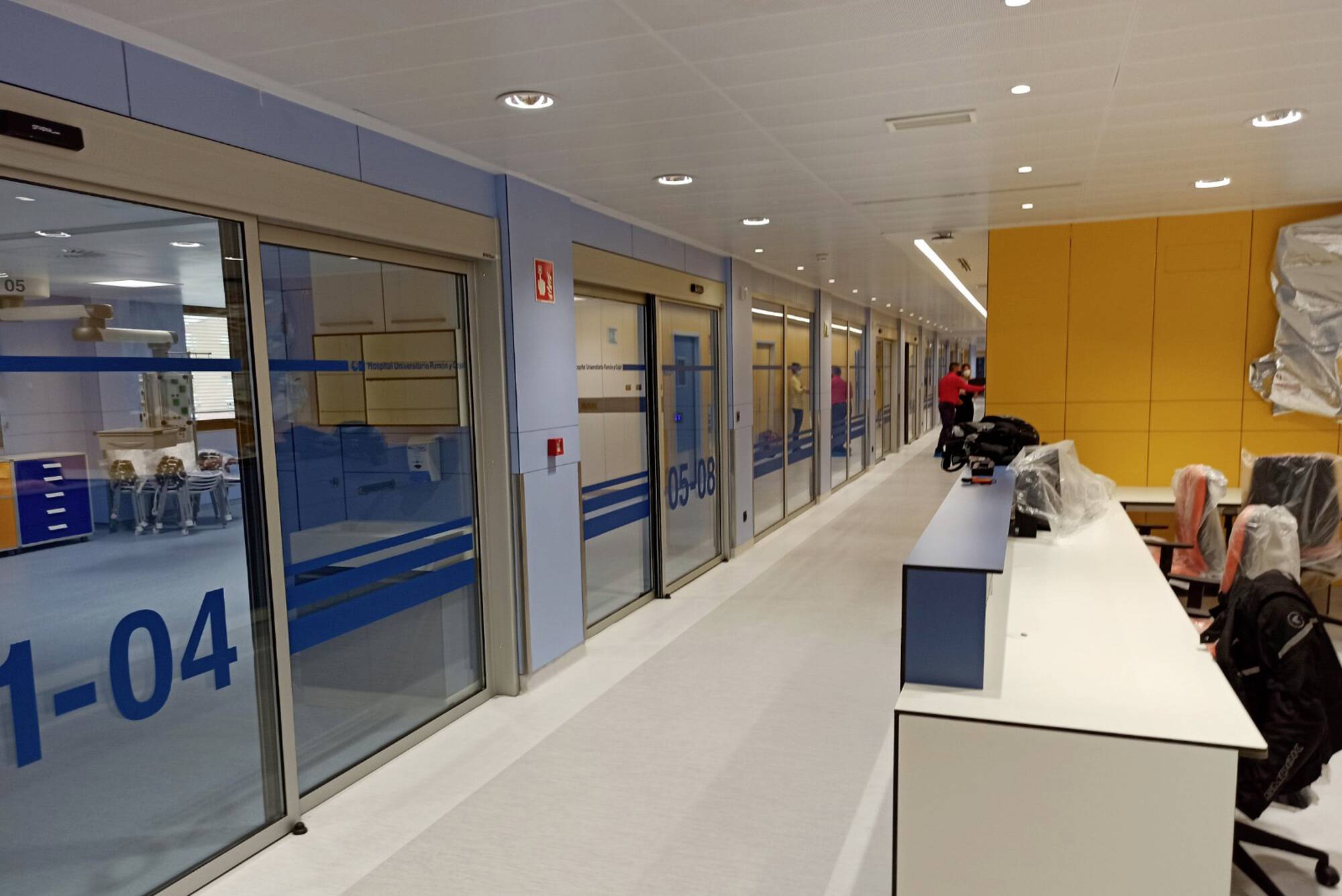 Ein moderner Krankenhausflur mit Glaswänden und blauen Streifen. Links sind nummerierte Räume, rechts ein Empfangsbereich mit Stühlen. Eine Person steht am Ende des Flurs.