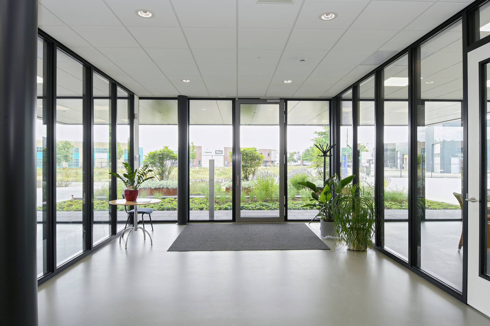 Eine moderne Eingangshalle mit großen Glaswänden, Pflanzen auf Tischen und einem Blick auf einen begrünten Außenbereich.