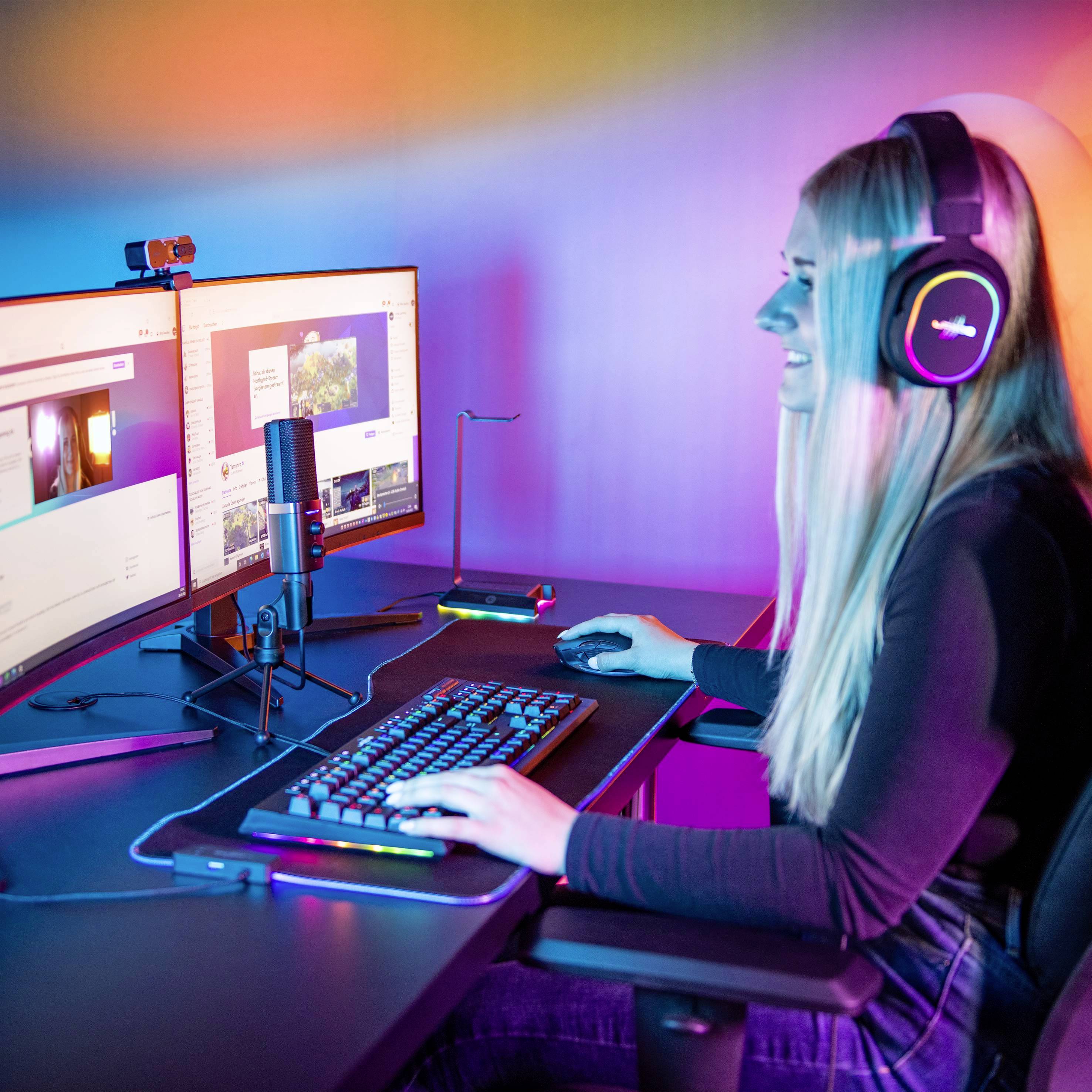 Eine Frau mit Kopfhörern sitzt an einem Computer mit zwei Monitoren. Sie arbeitet oder spielt, während sie in ein Mikrofon spricht.