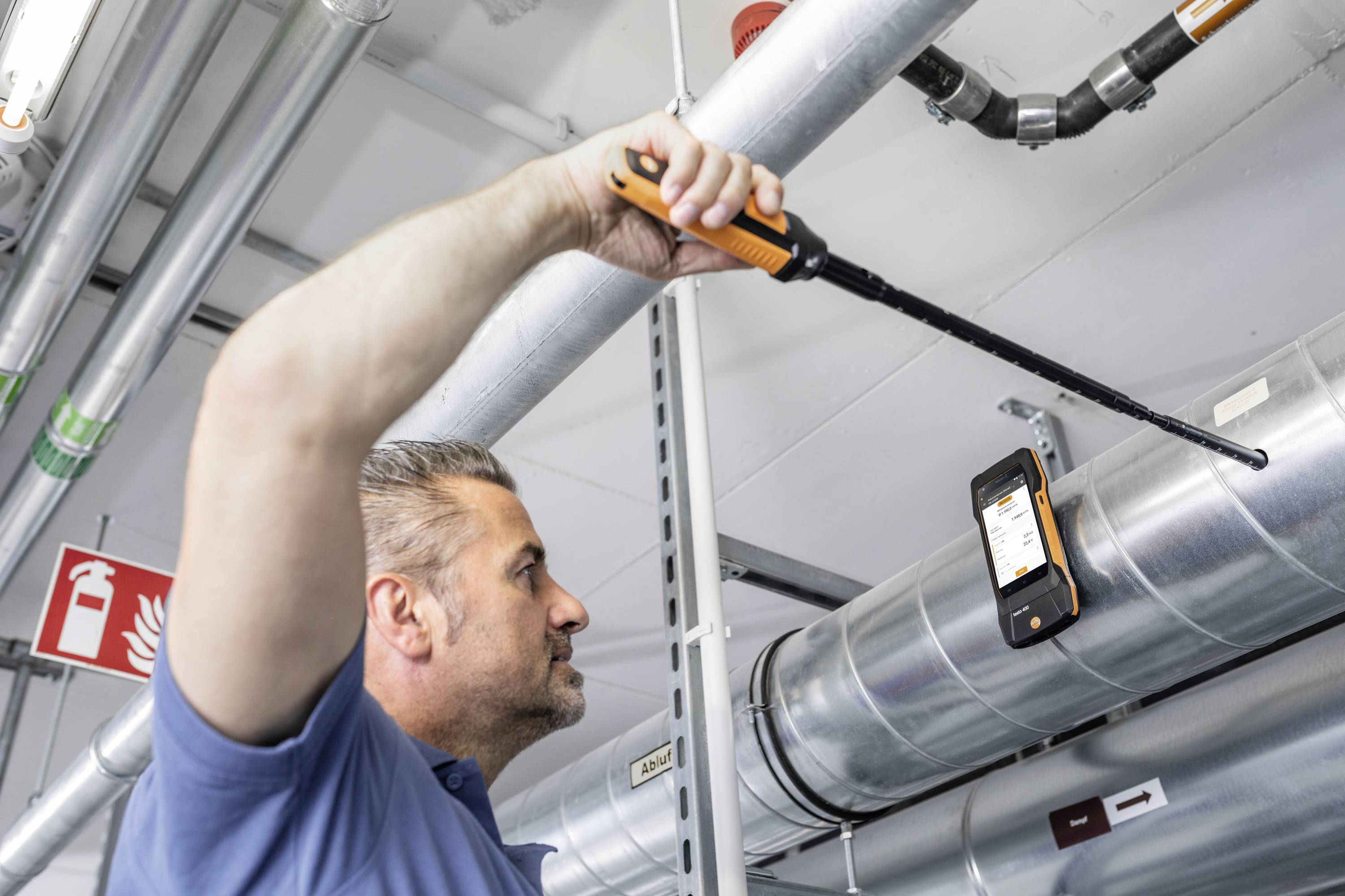 Ein Mann inspiziert ein Lüftungssystem mit einem langen Messgerät. Ein digitales Display zeigt Ergebnisse an. Umgebung industriell.