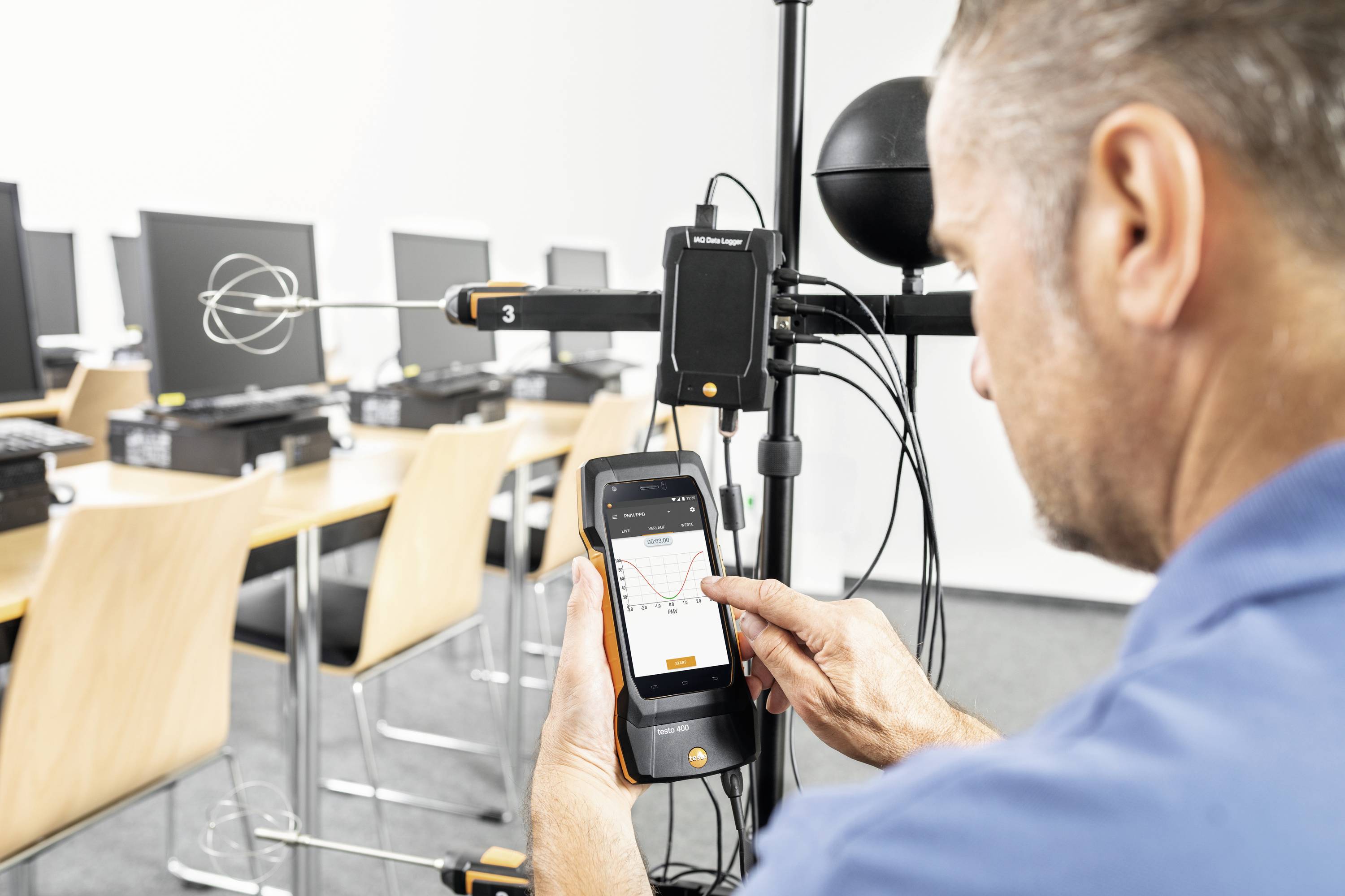 Ein Mann nutzt ein Messgerät in einem Raum mit Computern. Er betrachtet Daten auf einem mobilen Gerät, wobei Technik im Vordergrund steht.