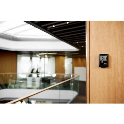 Ein Wandthermometer zeigt '54%' an, in einem modernen Bürogebäude mit Glasgeländern und Pflanzen im Hintergrund.