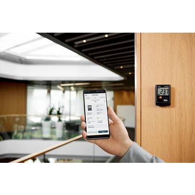 Eine Person hält ein Smartphone, das eine App zur Raumüberwachung zeigt, neben einem digitalen Thermostat auf 23,7 Grad Celsius eingestellt.