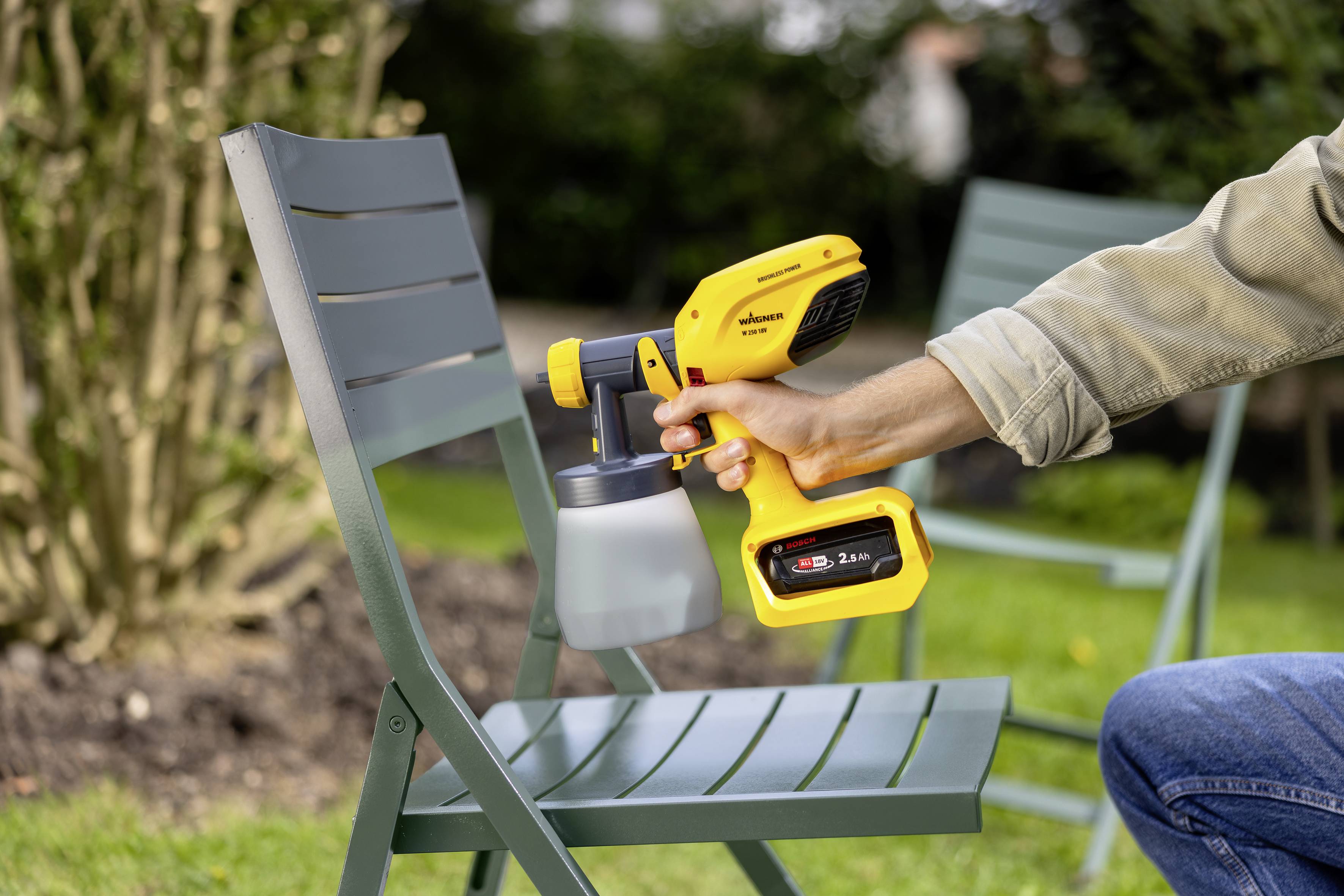Eine Person sprüht grüne Farbe auf einen Metallstuhl im Garten mit einem gelben Farbsprühgerät.