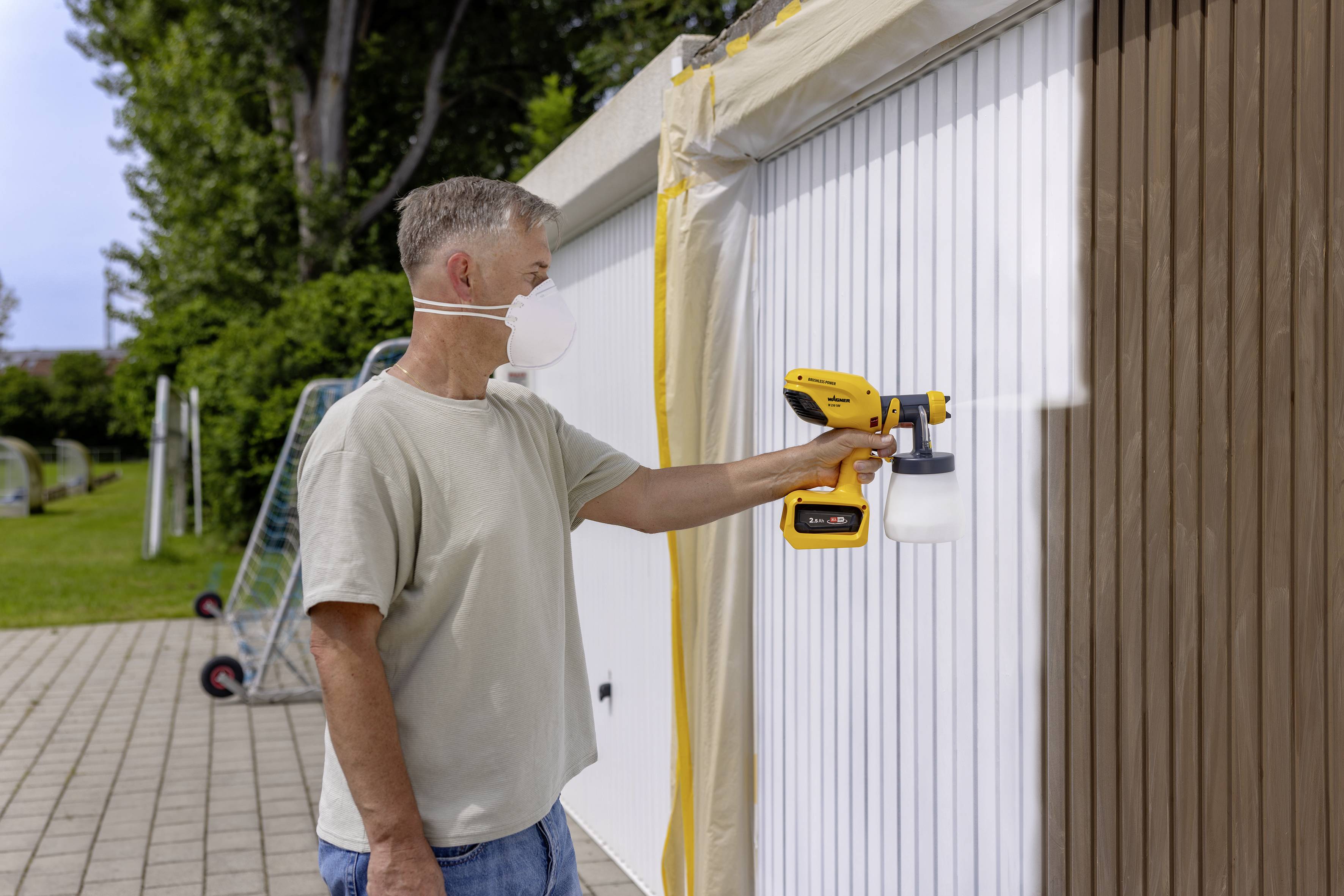 Ein Mann trägt eine Schutzmaske und sprüht Farbe mit einer Spritzpistole auf ein Garagentor im Freien.