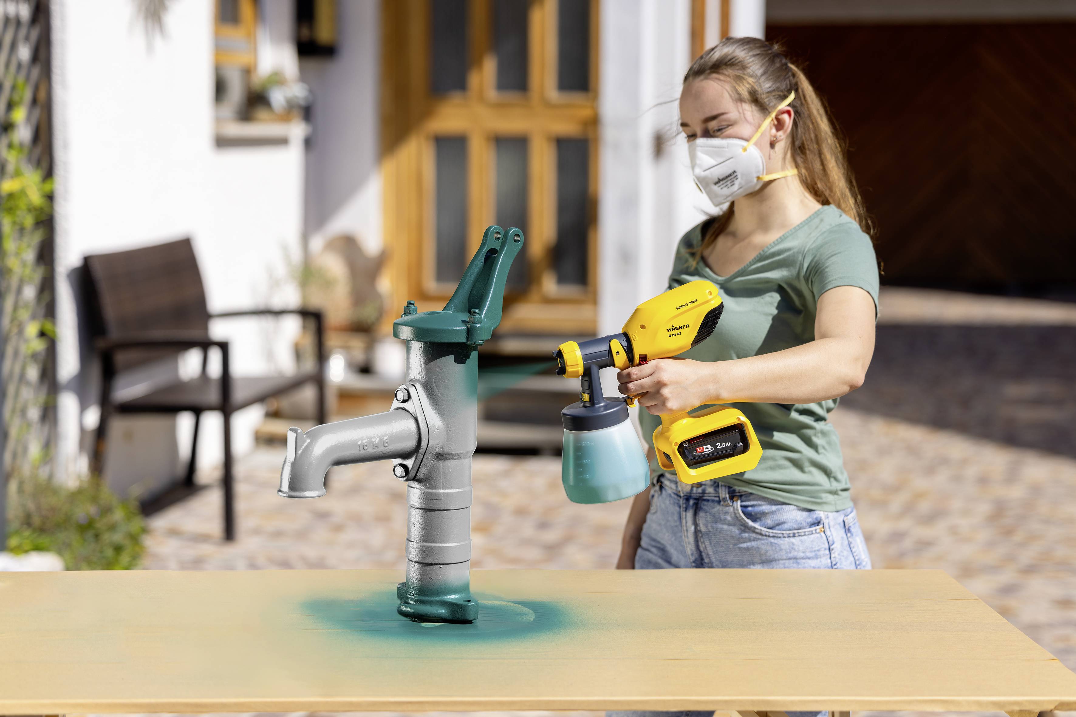 Eine Frau sprüht mit einer gelben Farbpistole eine grüne Farbe auf eine Metallpumpe. Sie trägt eine Schutzmaske und steht im Freien.