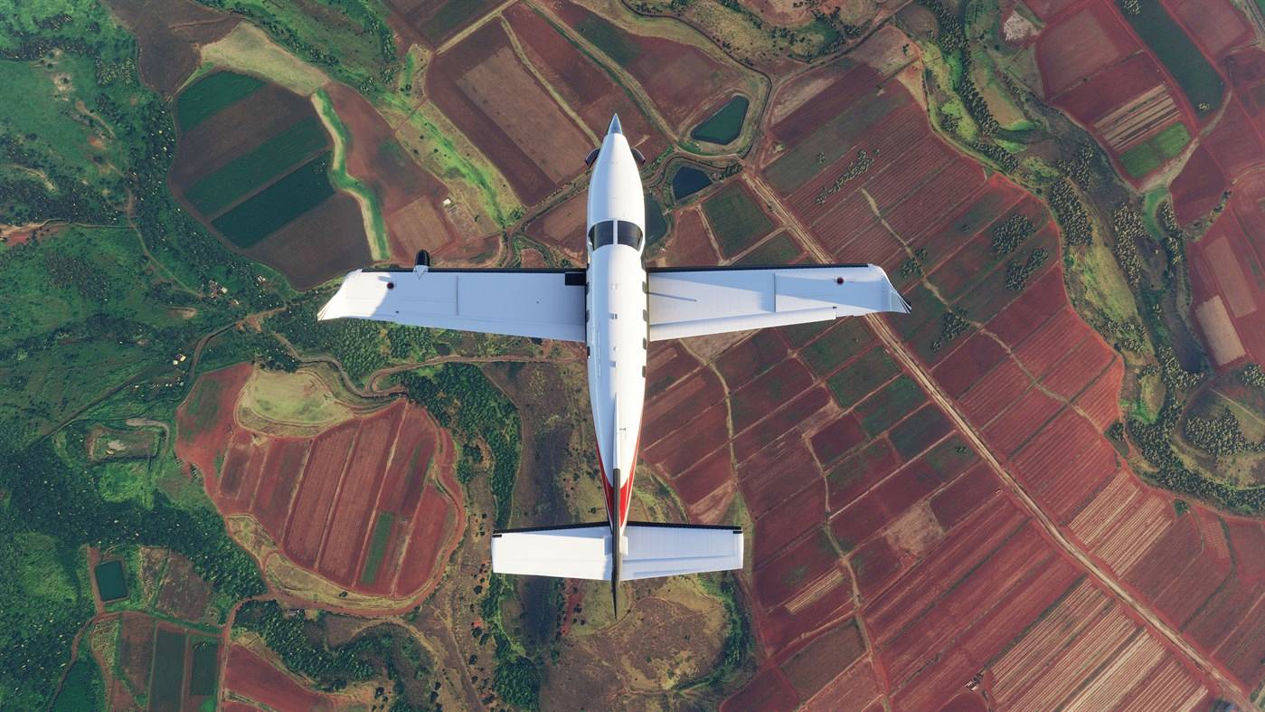Ein Flugzeug fliegt über bunte, gemusterte Felder. Die Landschaft zeigt unterschiedliche Brauntöne und Grünflächen aus der Vogelperspektive.