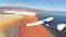 Ein Flugzeug fliegt über bunte Salzfelder, die in leuchtenden Rot- und Orangetönen schimmern. Ein weiter Blick über die Landschaft.