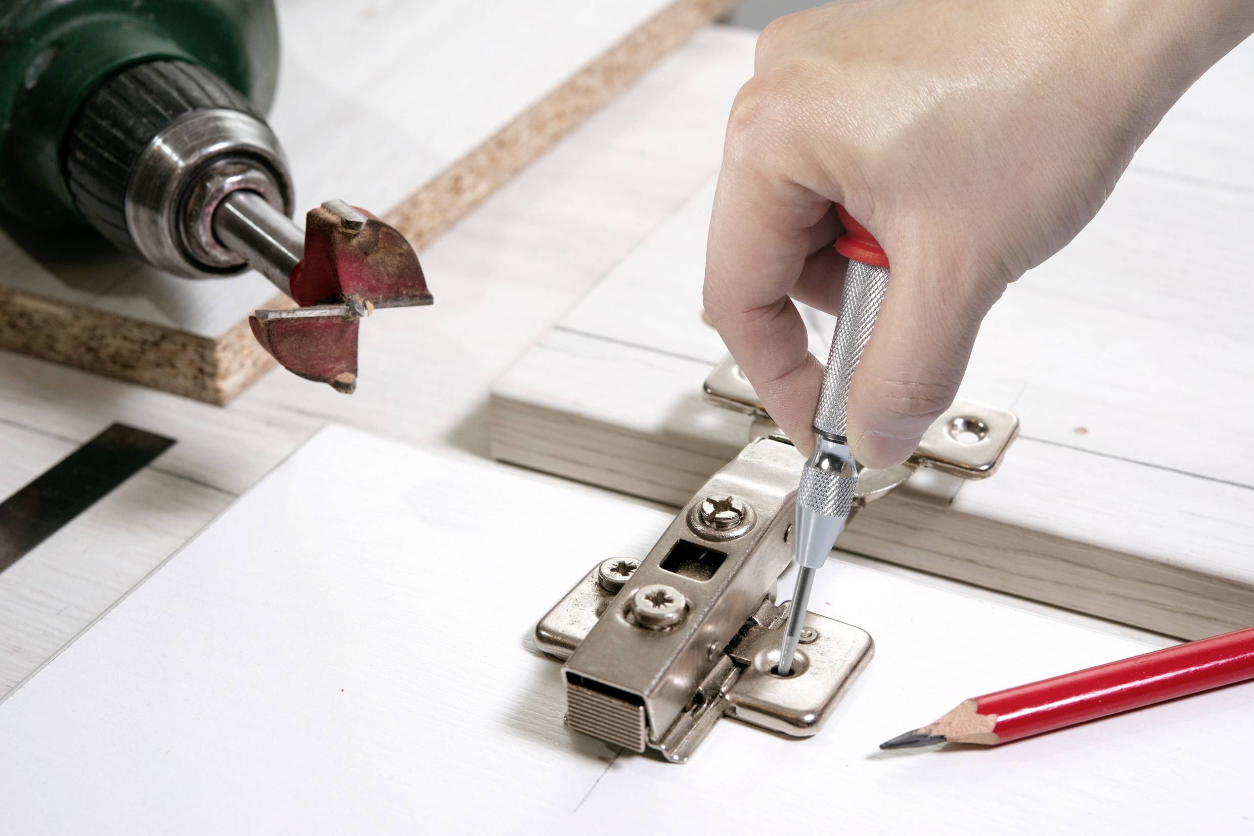 Eine Person befestigt ein Scharnier an einer Holzplatte mit einem Schraubendreher. Im Hintergrund liegt ein Bohrer.