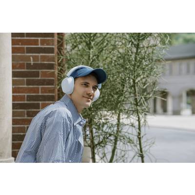 Ein Mann mit blauer Kappe und Kopfhörern sitzt draußen neben einer Pflanze und schaut zur Seite, im Hintergrund sind Gebäude sichtbar.