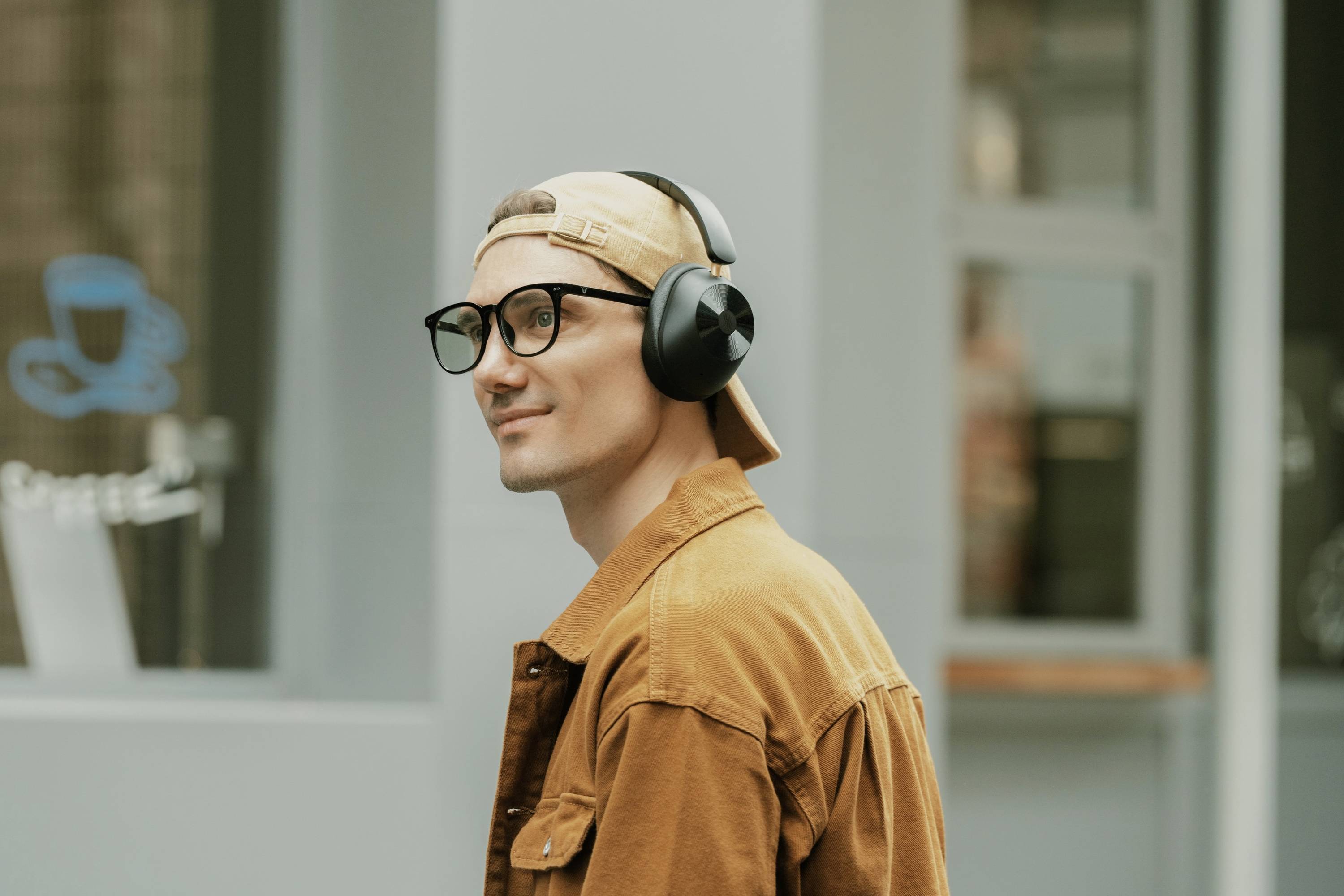 Eine Person mit Brille und Kopfhörern geht entspannt draußen, trägt eine braune Jacke und eine beige Mütze rückwärts.