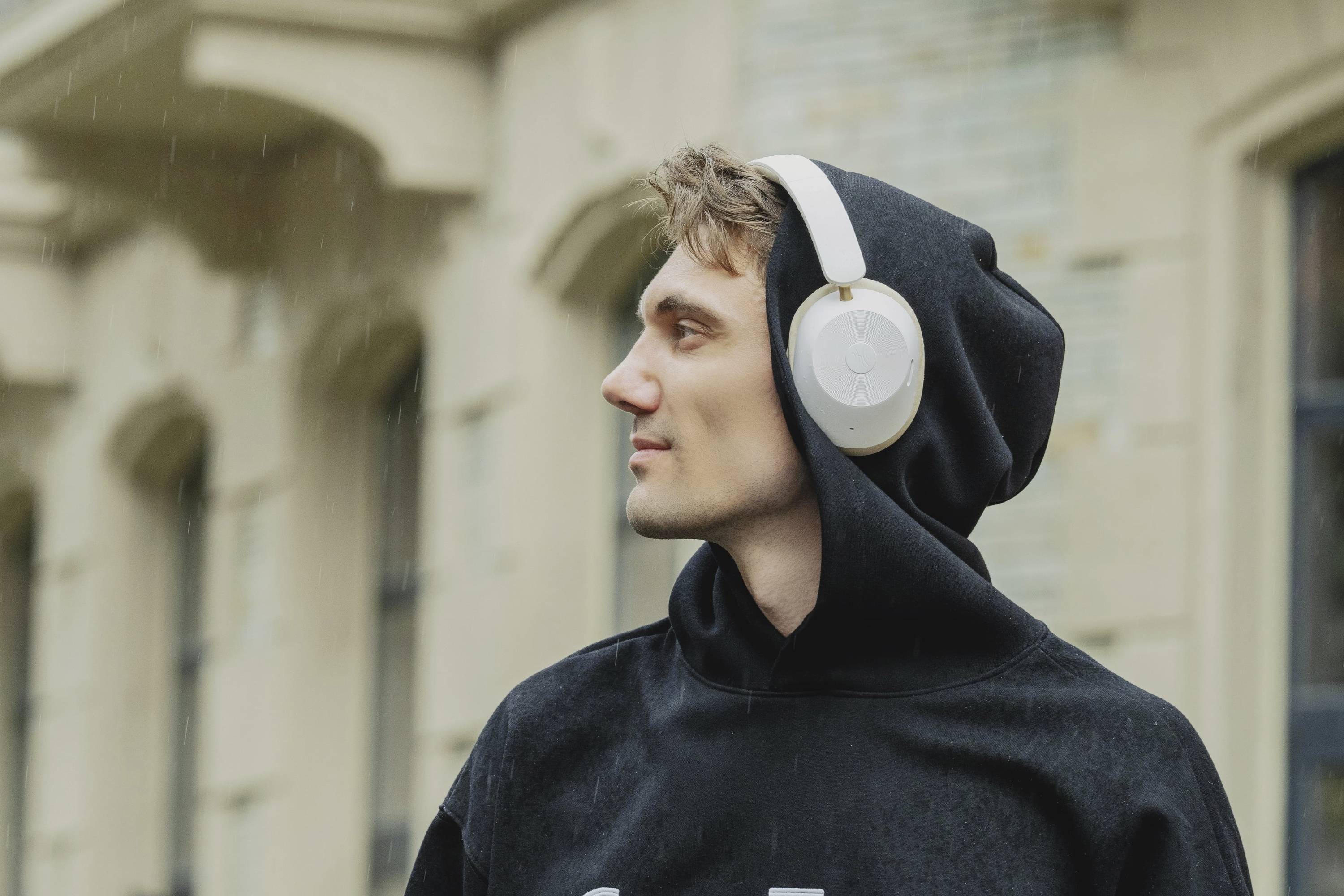 Eine Person mit Kapuze hört Musik über Kopfhörer und schaut nach rechts. Im Hintergrund sind Gebäude zu sehen.