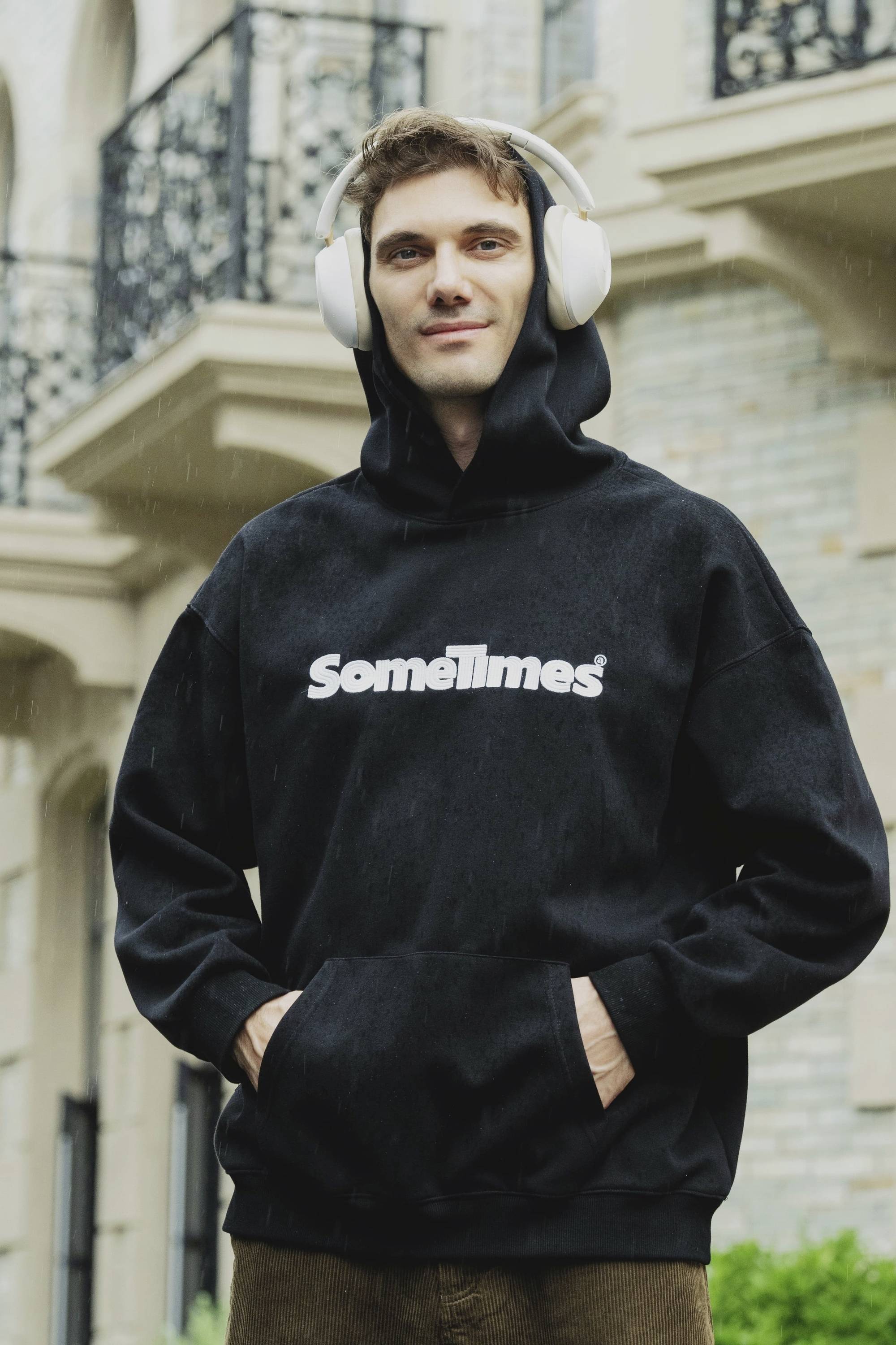 Ein Mann trägt Kopfhörer und einen schwarzen Hoodie mit dem Aufdruck 'Sometimes'. Er steht vor einem Gebäude mit Balkonen.