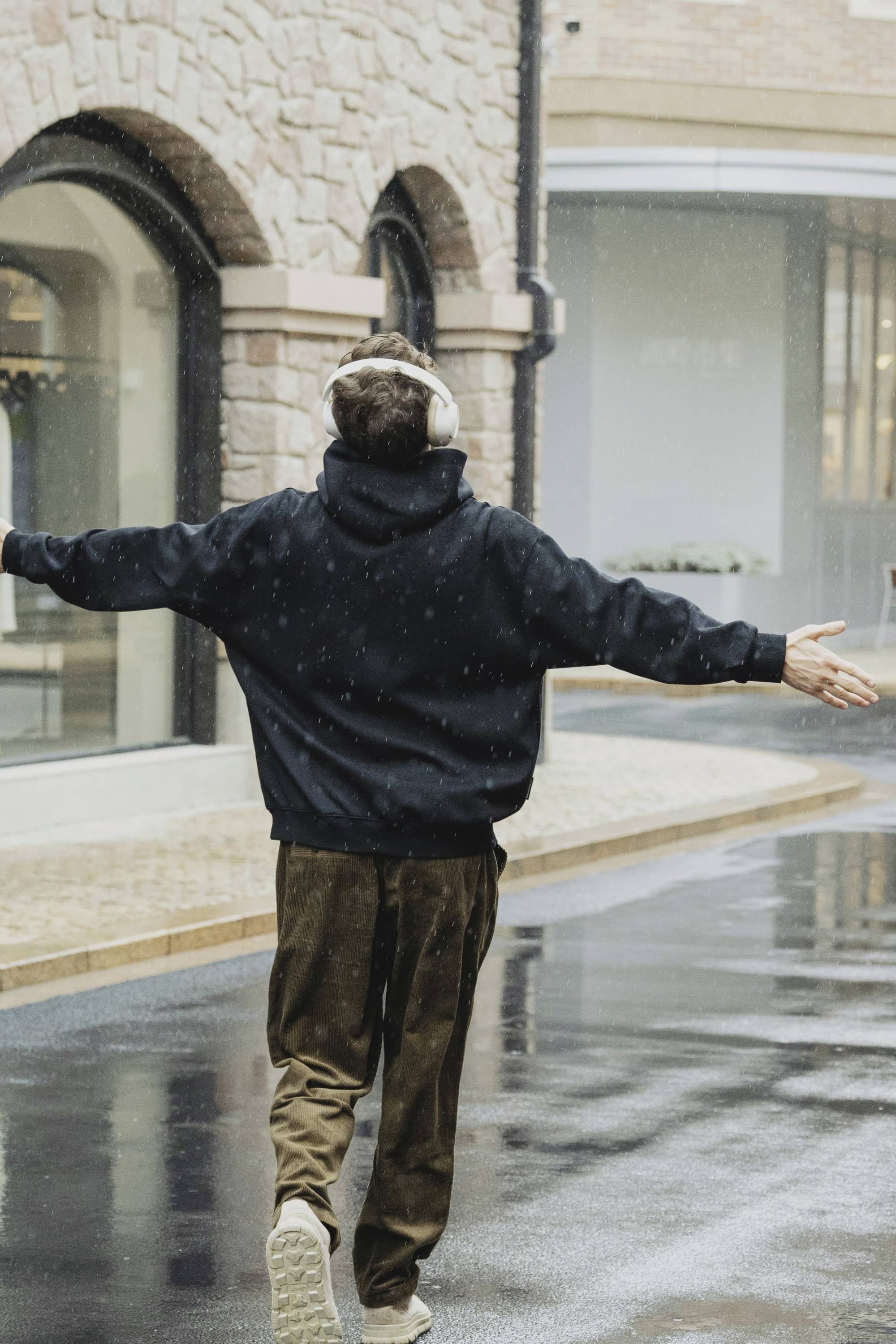 Person mit Kopfhörern geht fröhlich auf einer nassen, gepflasterten Straße, Arme ausgebreitet, während es regnet.