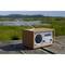 Ein Retro-Radio aus Holz steht auf einem Holztisch im Freien. Im Hintergrund sind unscharf Graslandschaft und Hügel zu sehen.