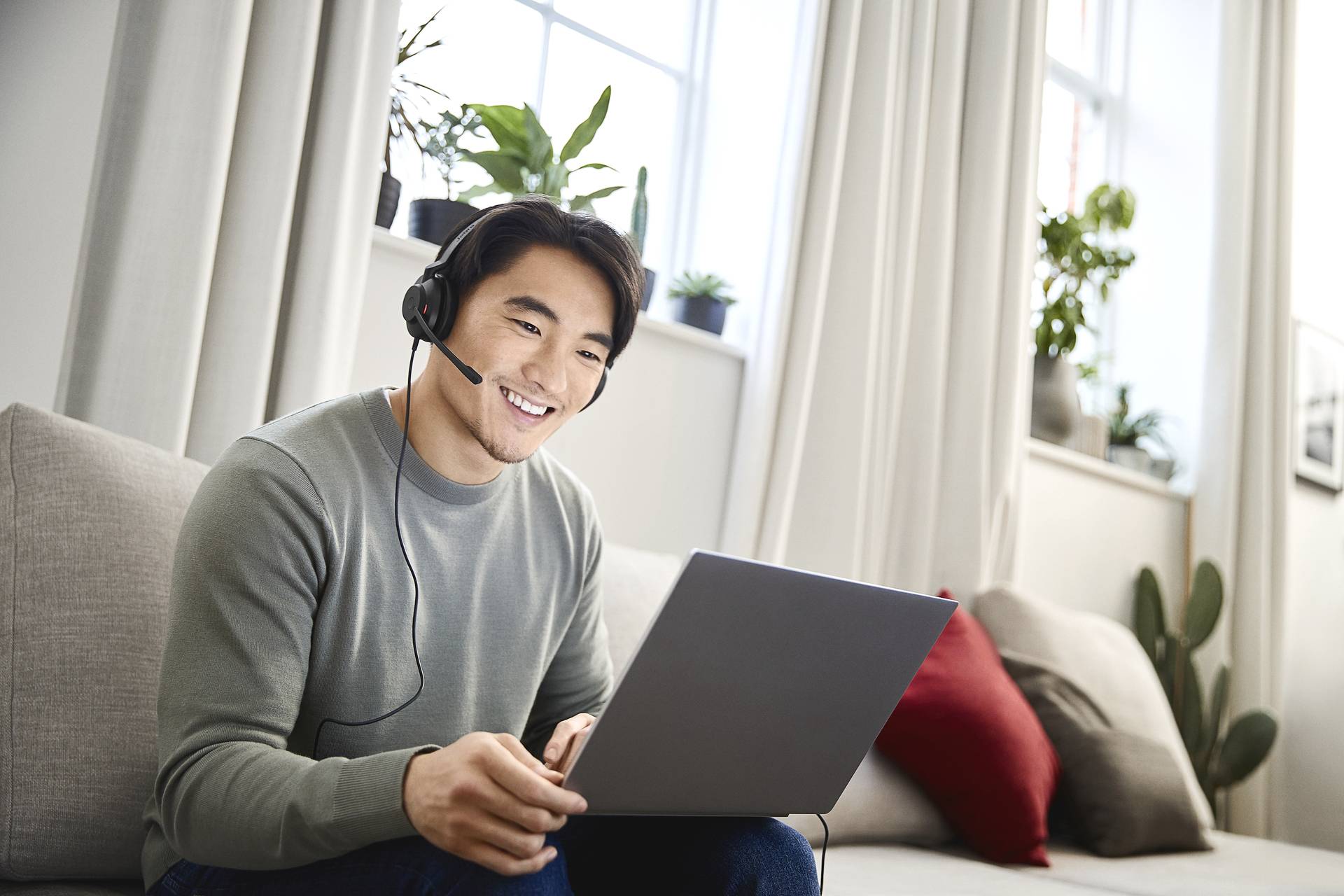 Ein lächelnder Mann mit Headset sitzt auf einem Sofa und benutzt einen Laptop. Im Hintergrund sind helle Fenster und Zimmerpflanzen zu sehen.
