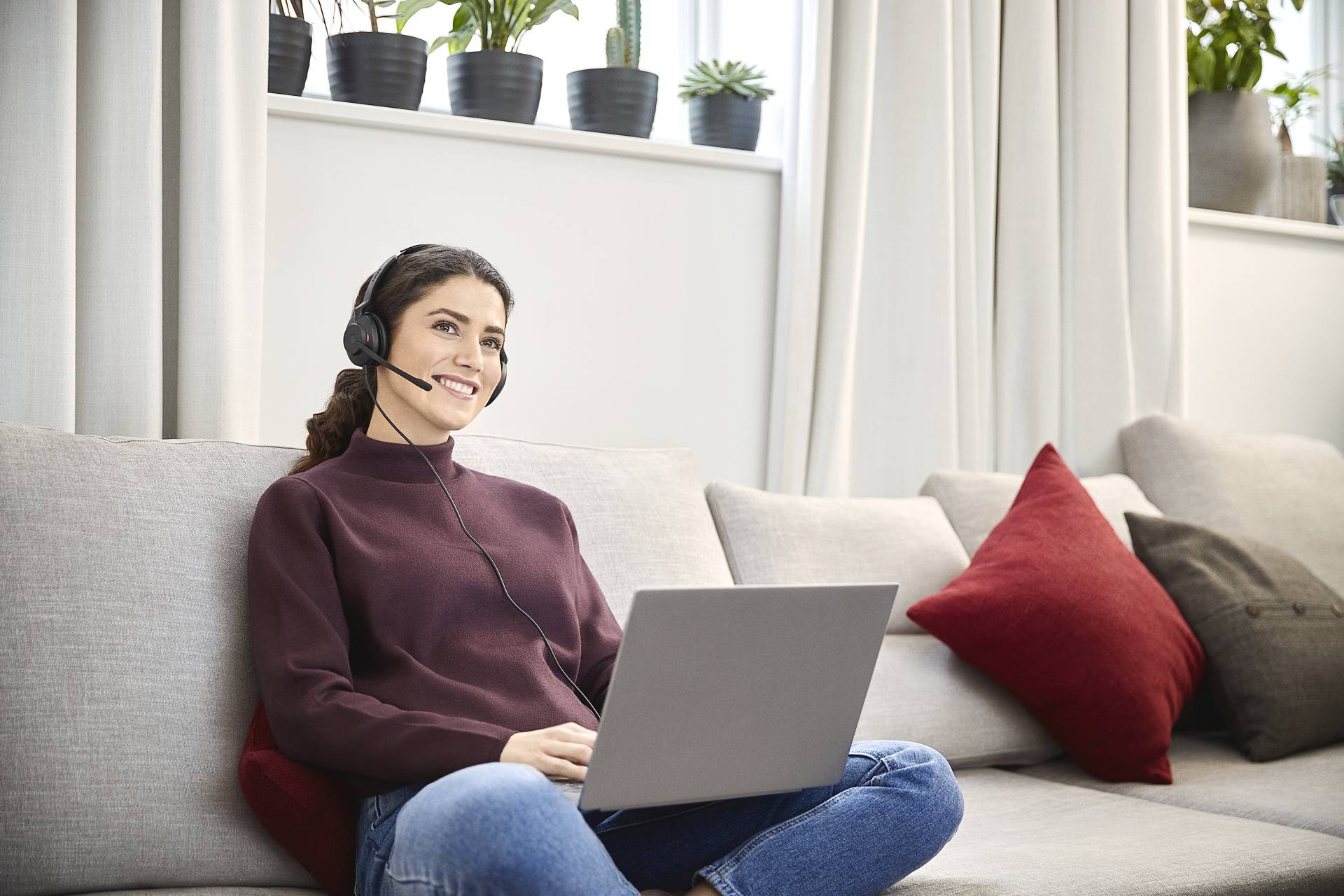 Frau mit Headset arbeitet lächelnd am Laptop auf dem Sofa, umgeben von Zimmerpflanzen auf einer Fensterbank.