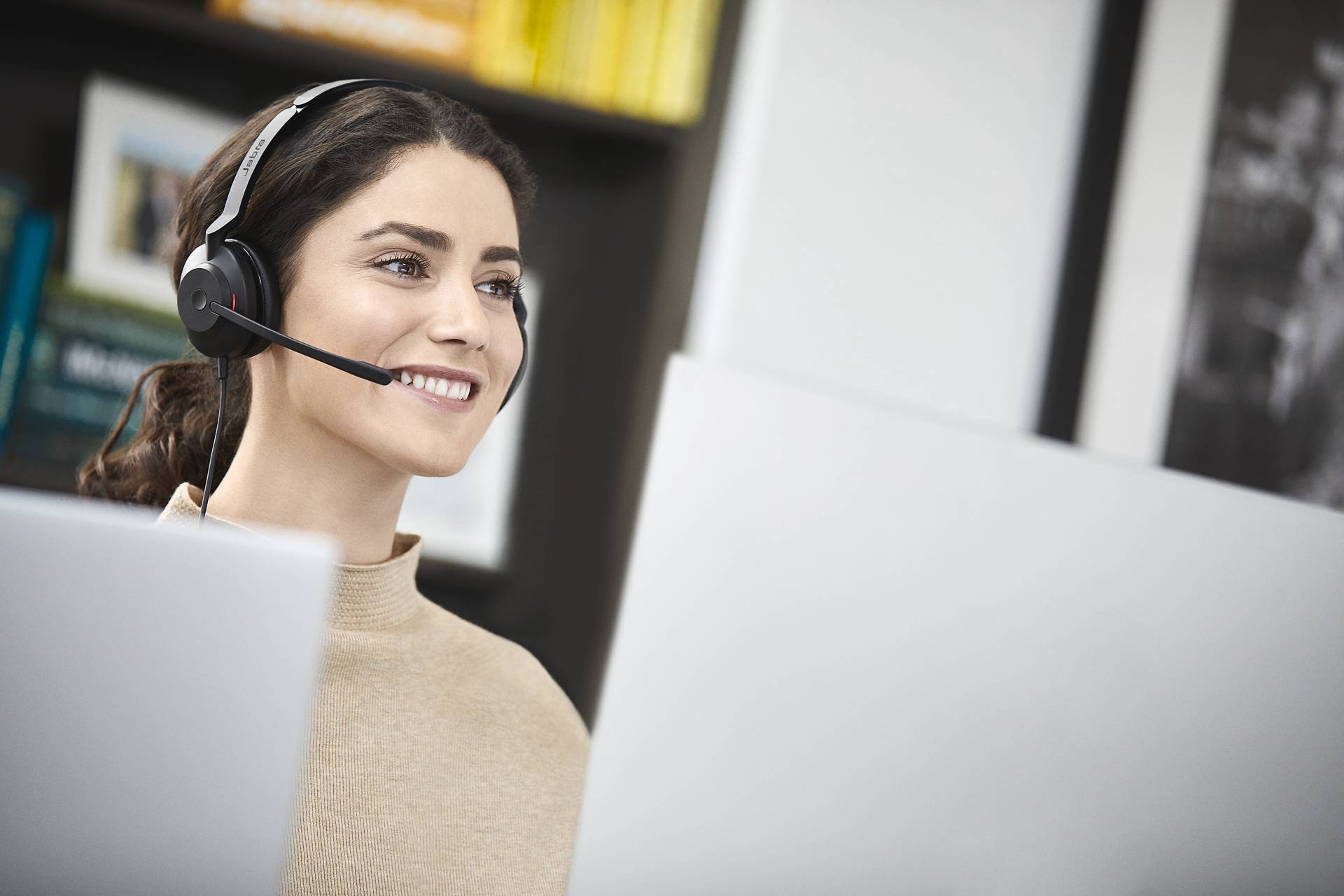Eine lächelnde Frau mit Headset sitzt vor einem Computerbildschirm, arbeitet möglicherweise in einem Callcenter oder Büro.