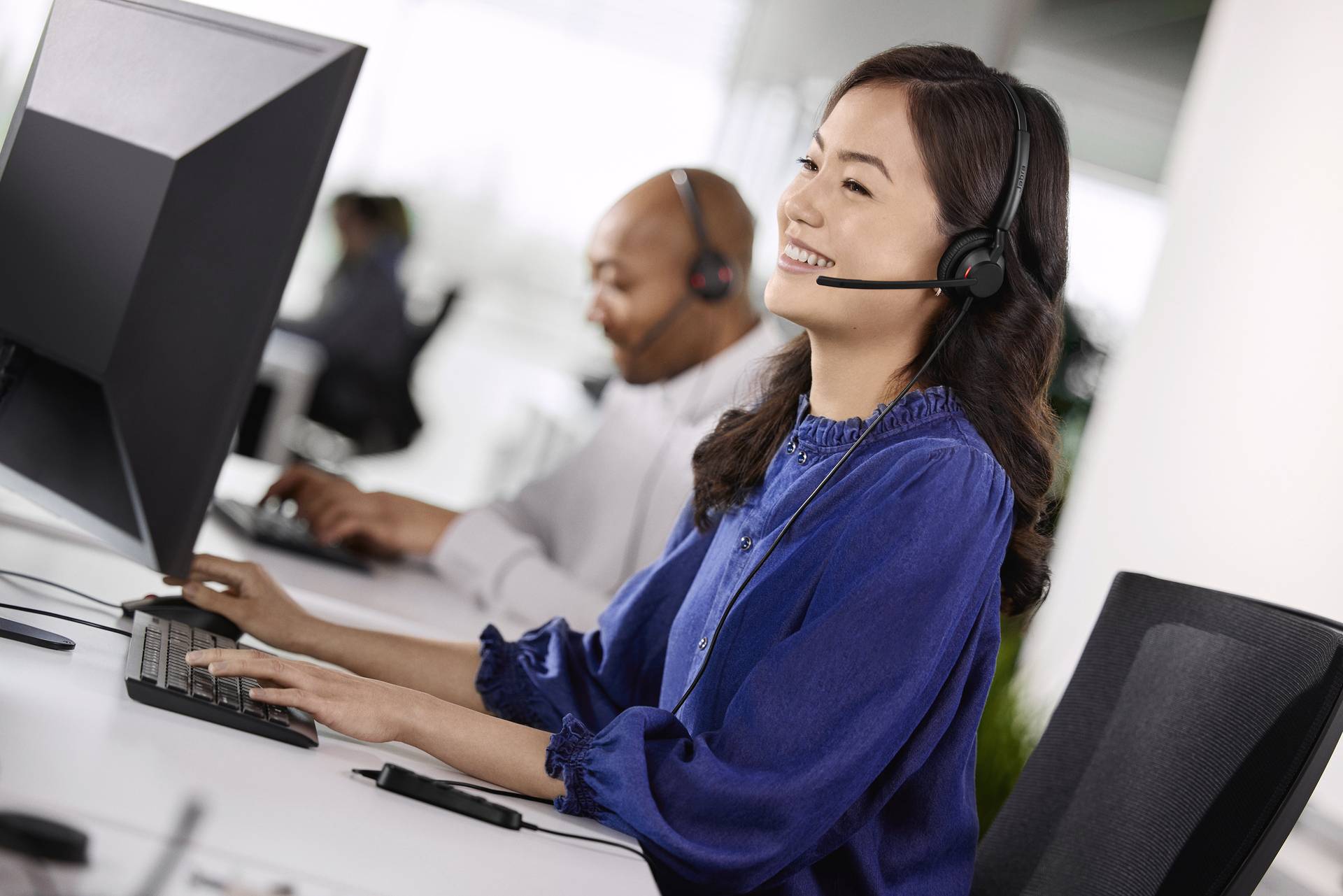 Eine lächelnde Frau mit Headset arbeitet am Computer, während ein Mann im Hintergrund ebenfalls am Computer arbeitet.