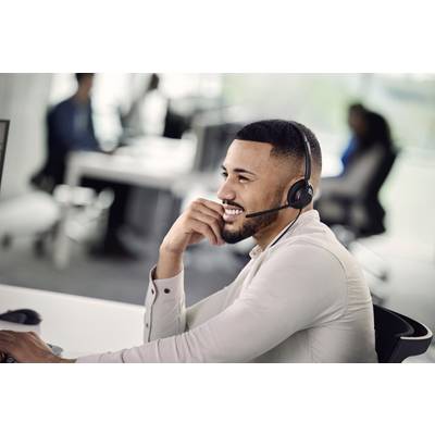 Mann mit Headset arbeitet lächelnd am Computer in einem Büro. Im Hintergrund sind unscharf weitere Personen an Schreibtischen zu sehen.