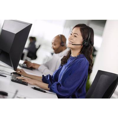 Eine lächelnde Frau trägt ein Headset und arbeitet am Computer, im Hintergrund ein weiterer Mitarbeiter. Büroarbeitsplatz-Szene.