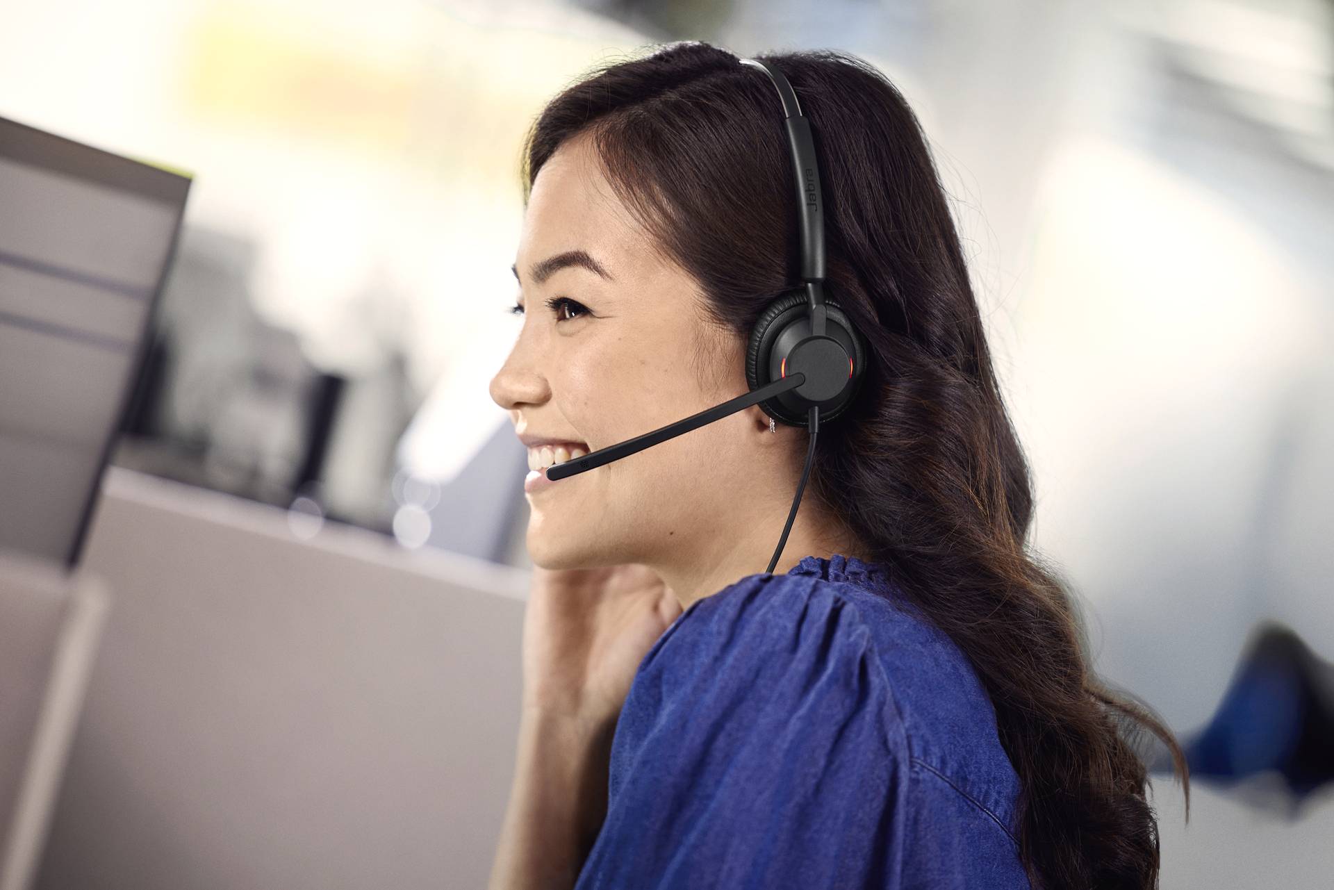 Frau mit Headset sitzt lächelnd vor einem Computerbildschirm, spricht mit einem Kunden. Büroumgebung im Hintergrund.