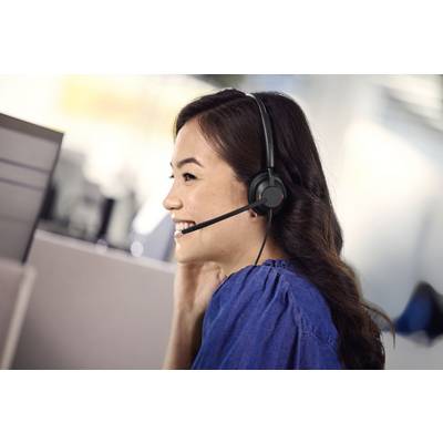 Frau mit Headset sitzt lächelnd vor einem Computerbildschirm, spricht mit einem Kunden. Büroumgebung im Hintergrund.