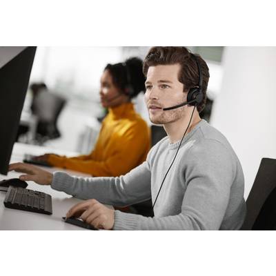 Ein Mann mit Headset sitzt an einem Computer und spricht, wahrscheinlich in einem Callcenter. Im Hintergrund ist eine weitere Person zu sehen.