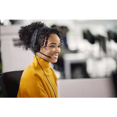 Eine Person mit einem Headset sitzt an einem Schreibtisch und lächelt. Der Hintergrund zeigt ein geschäftiges Büro.