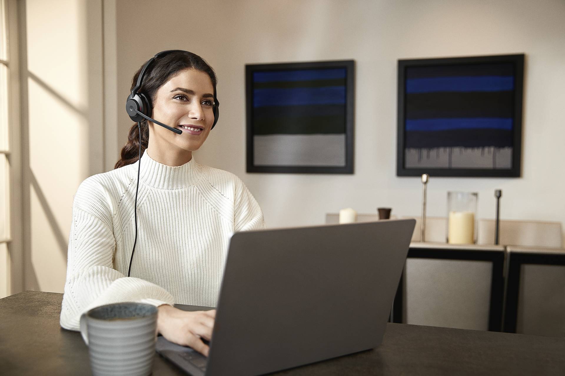 Eine Frau sitzt mit Headset vor einem Laptop, lächelt und arbeitet wahrscheinlich. Im Hintergrund sind gerahmte Bilder an der Wand.