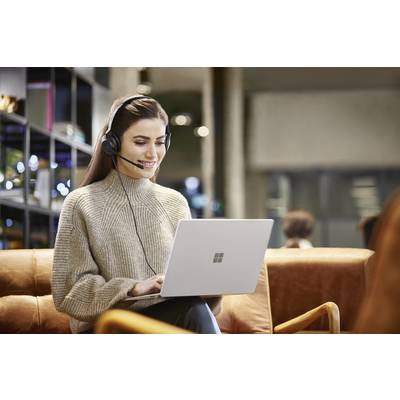 Eine Frau mit Kopfhörer sitzt auf einem Ledersofa und arbeitet an einem Laptop, lächelnd. Sie ist in einer modernen Arbeitsumgebung.