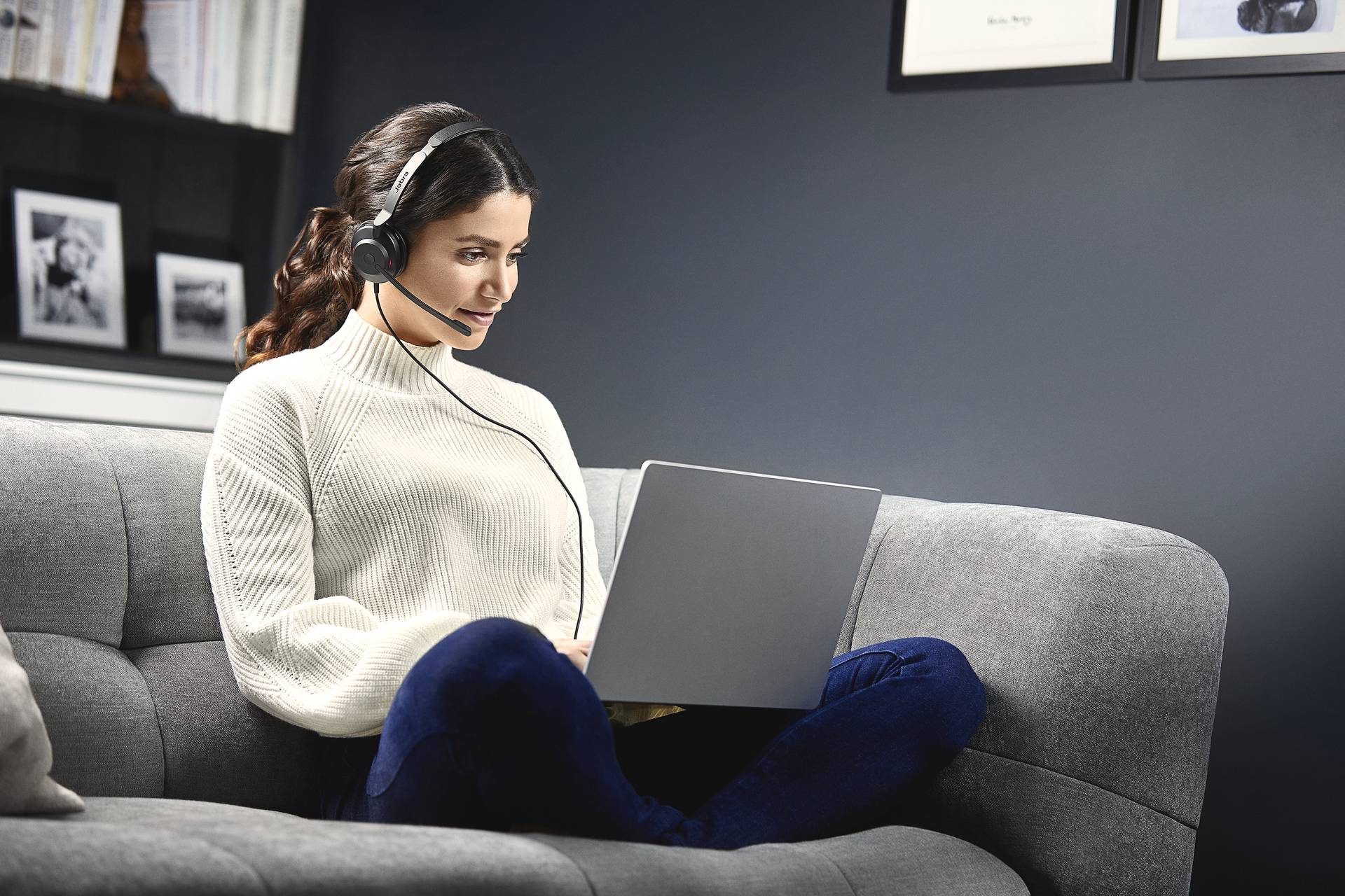 Eine Frau sitzt auf einem grauen Sofa, trägt ein Headset und arbeitet an einem Laptop. Im Hintergrund sind Bücherregale zu sehen.